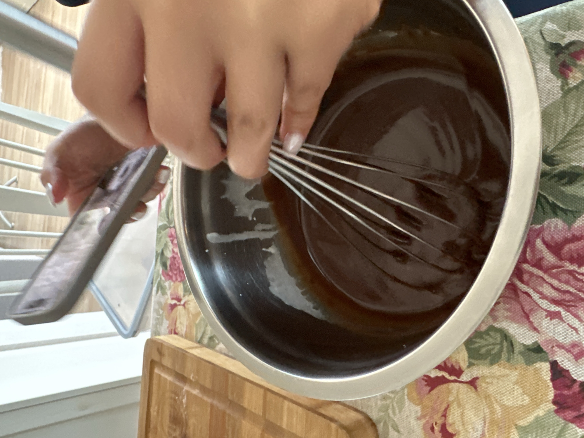 A close-up, eye-level shot captures a person whisking a dark brown substance in a stainless steel bowl, likely a cooking activity. The person's hands are visible, one holding the whisk and the other holding a smartphone with a mirrored image on the screen. The bowl sits on a wooden cutting board atop a floral-patterned tablecloth. In the background, a white metal staircase with horizontal bars is partially visible. Without further information, the location cannot be identified in Varna, Bulgaria.