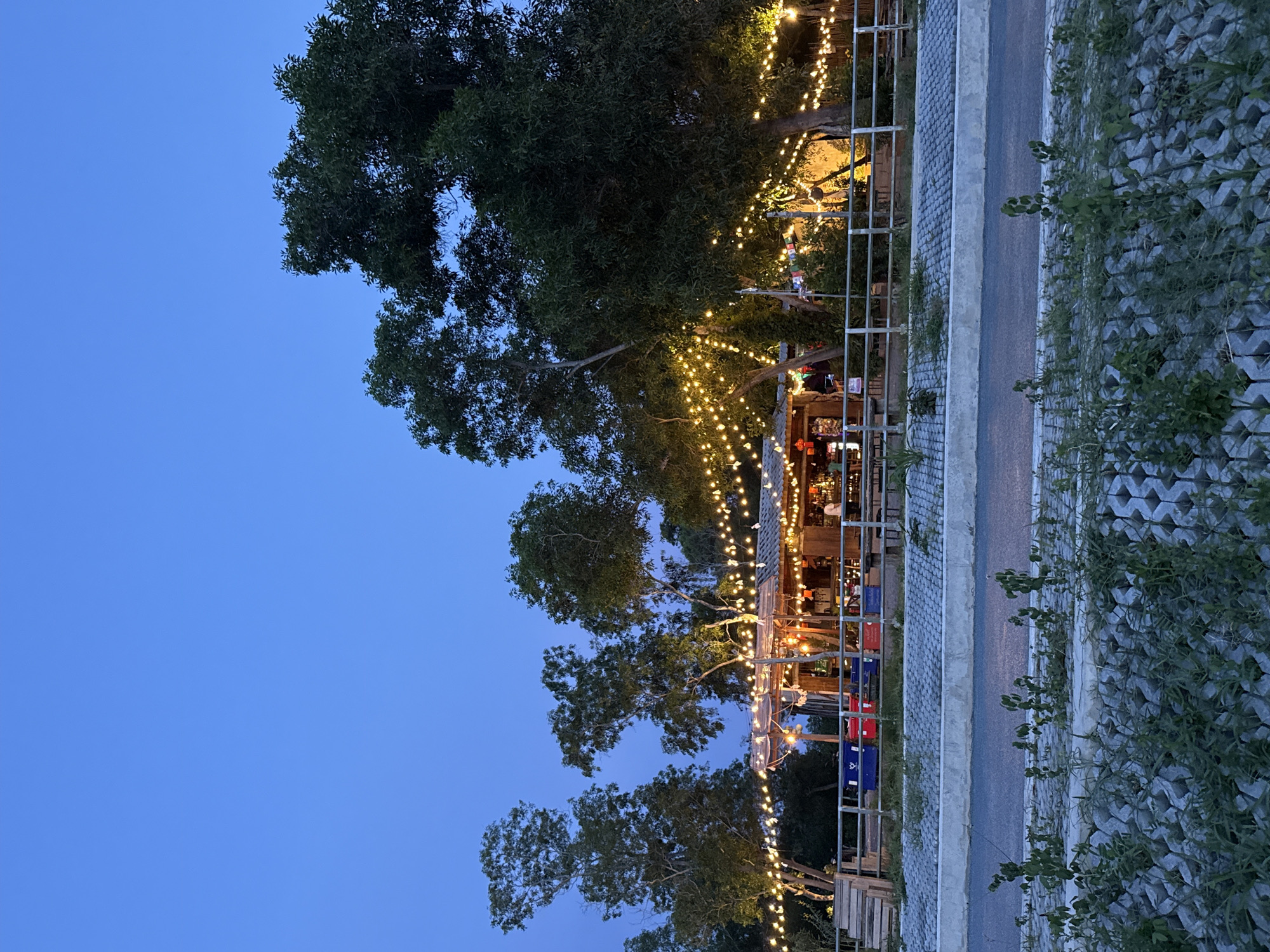The image depicts a bar or restaurant at dusk in Baan Chaweng, Thailand. String lights are illuminated, casting a warm glow over the open-air structure, which appears to be a wooden building with a corrugated metal roof. Several trees with dense foliage surround the establishment. In the foreground, a paved walkway with a concrete border runs parallel to a grey road. Beyond the road, there is a rocky embankment covered with sparse green vegetation. A metal railing separates the walkway from a seating area in front of the bar. Several blue and red crates are visible behind the railing. The sky is a deep blue, indicating twilight.