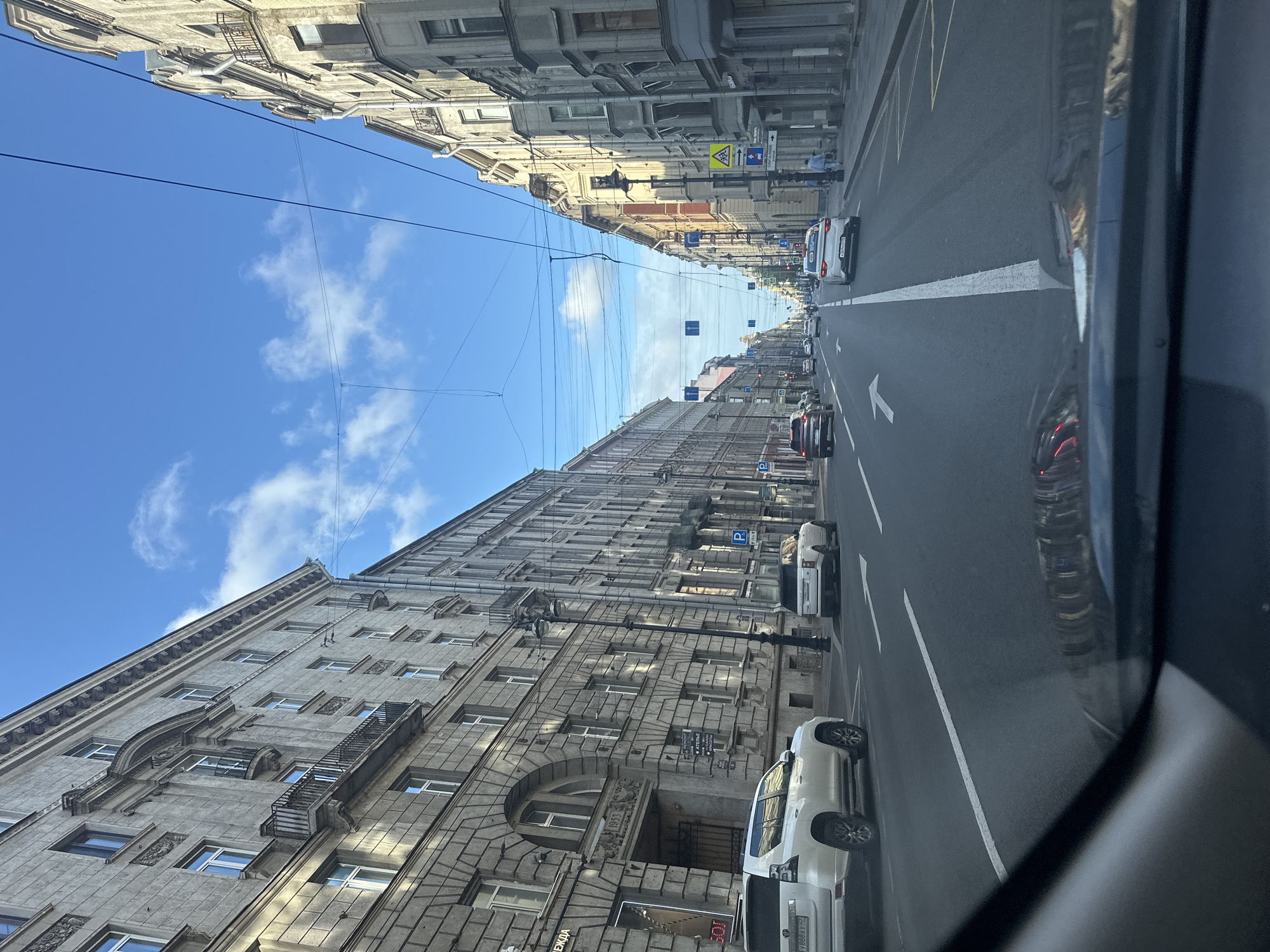 The image depicts a street scene in Saint Petersburg, Russia, viewed from the perspective of a vehicle moving forward. The street is lined with tall, multi-story buildings, characteristic of the city's architecture, with ornate facades and numerous windows. The sky is blue with scattered white clouds.
Several cars are visible on the road, including a white SUV on the left, a white Range Rover on the right, and other vehicles further down the street. Traffic lights and street signs are present. The road has clear lane markings. Power lines are strung across the street above the buildings. On the left side of the street, there is a building with "1953" inscribed above an archway, and other buildings have shops or businesses on the ground floor, indicated by signage. Pedestrians are visible walking on the sidewalk on the right side of the street. The overall impression is of a typical urban environment in Saint Petersburg on a clear day.