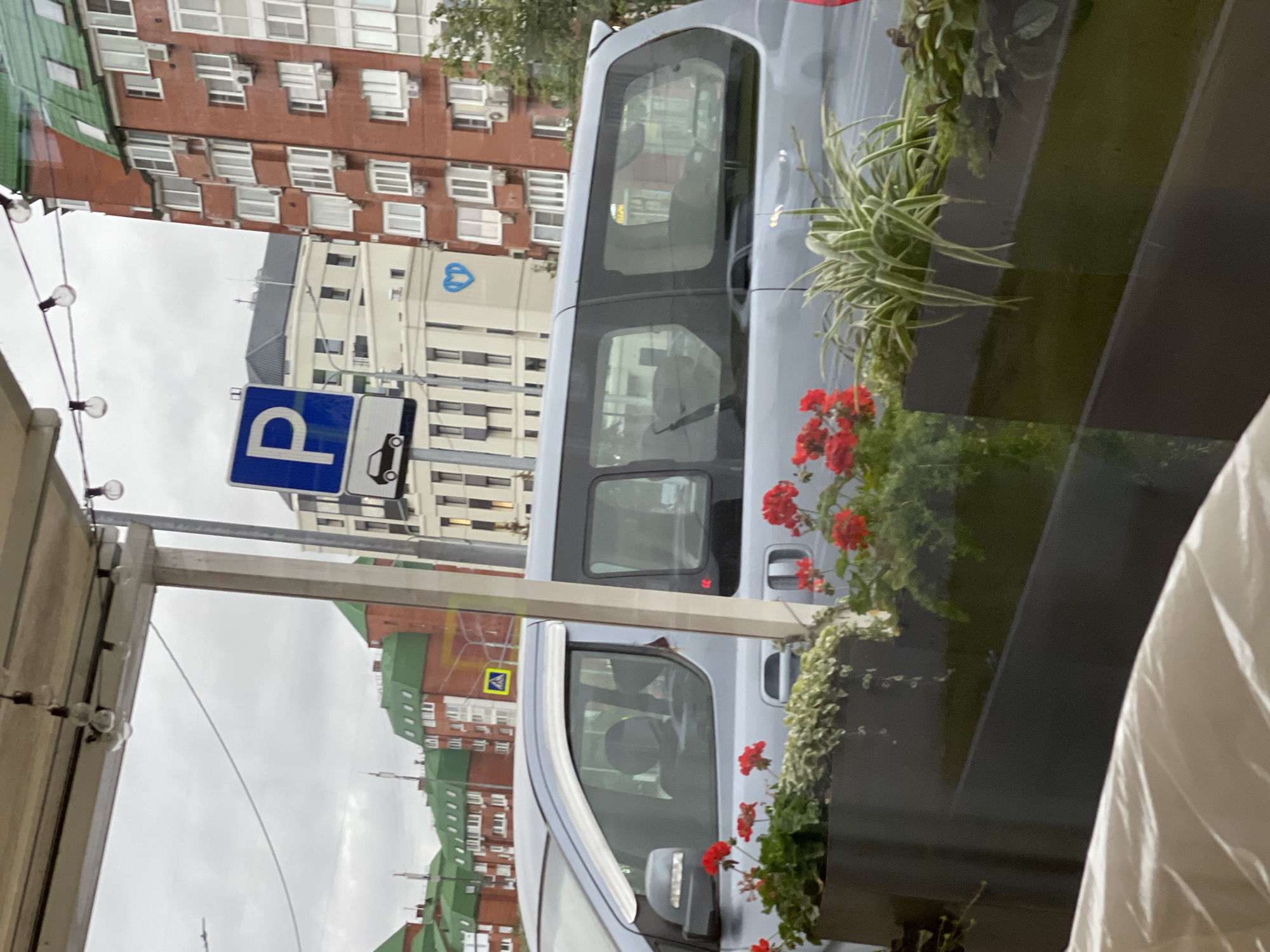 The image captures a scene from Moscow, Russia, featuring a light blue minivan parked near a street with several multi-story buildings. A prominent blue parking sign with a white 'P' and a silhouette of a car is visible. In the foreground, a dark planter box holds red geraniums and spider plants. The buildings in the background are a mix of red brick structures with green roofs and lighter colored buildings. One of the lighter buildings displays a blue heart-shaped logo. The sky is overcast, suggesting a cloudy day. The viewpoint appears to be from an elevated position, possibly a cafe or balcony, looking outwards towards the street.