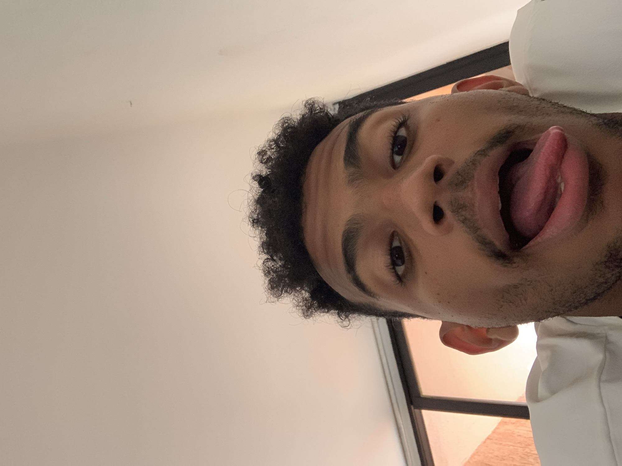 The media file features a close-up portrait of a young man, rotated 90 degrees counter-clockwise. He has dark, curly hair, dark brown eyes, and stubble or short facial hair. The man is looking directly at the camera with a playful and humorous expression, sticking his tongue out. His mouth is wide open, revealing his tongue and teeth. A small part of his ear is visible on the right side of his face.
He appears to be indoors, possibly lying down, with the camera positioned above him. The background is predominantly a plain, light cream or off-white surface, likely a wall or ceiling. On the right side of the frame, behind his head and shoulder, a window with a dark frame is visible. Through the window, an orange-hued light is apparent, suggesting either a warm indoor light source, or the natural light of a sunrise or sunset outside. A part of a white garment or pillow is also visible on the far right, near his shoulder.
The overall mood is lighthearted and informal. There is no visible text in the image. The implied location context for this image is Porlamar, Venezuela.