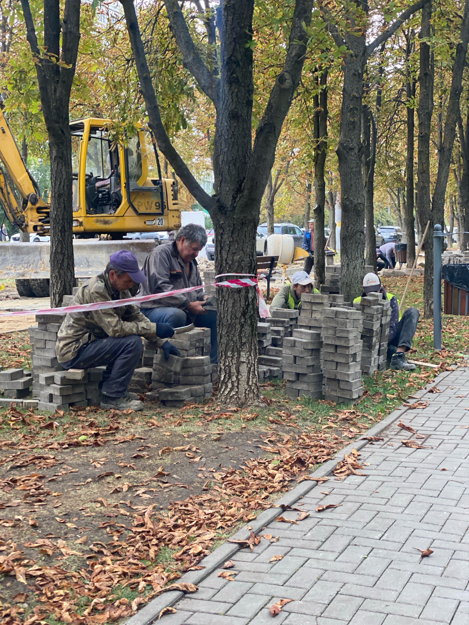 In Chișinău, Moldova, a construction scene unfolds in an autumnal environment, with a yellow excavator visible in the background. Several men are engaged in construction work, with stacks of gray paving stones piled around them. Two men are seated on stacks of stones, one wearing a purple cap and a camouflage jacket, the other in a gray jacket. Another man in a yellow vest and white cap is also working with the stones, and a fourth man in a yellow vest and cap is seated nearby. A red and white striped construction tape is visible, suggesting a work area. Fallen brown leaves litter the ground around the trees, indicating the season. A paved walkway runs along the right side of the image.