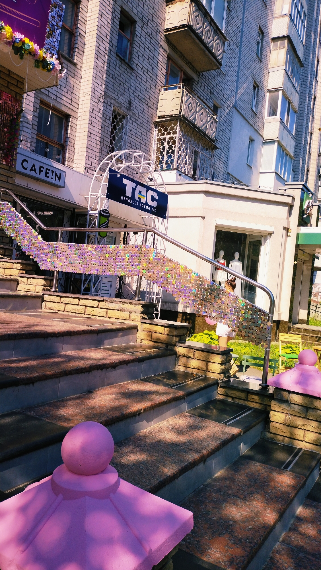 The image captures an exterior view of businesses located in the city of Horishni Plavni, Ukraine. The foreground features a set of granite-clad outdoor stairs, with some steps exhibiting a speckled reddish-brown pattern. The stairs are flanked by brick structures and are decorated with a shimmering, iridescent sequin curtain draped over the handrails. Two bright pink, decorative finials are visible at the base of the stairs, adding a pop of color to the scene.
Further up, a person in a white top and skirt is standing near the entrance of a shop, holding a pink balloon. Behind the stairs, the facade of a building displays signage for "CAFE:N" and "TAC", with the latter having the text "страхова група ТАС" underneath. The building's brickwork is visible, along with several windows and balconies with decorative metal railings. The scene is bathed in sunlight, casting shadows across the stairs and highlighting the textures of the materials.