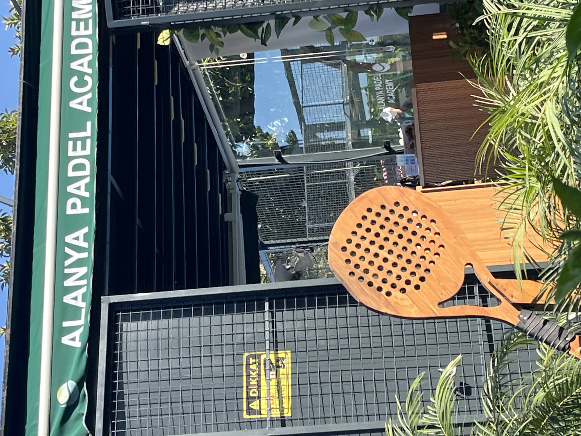The image shows a building identified as "Alanya Padel Academy," as evidenced by signage. A large, wooden paddle-like object with circular perforations is prominently displayed against a dark gray grid fence. A warning sign with the word "DIKKAT" (Turkish for "Attention") is visible on the fence. Green foliage is present on the right side of the image, partially obscuring the paddle and fence. The building's structure includes glass elements reflecting the sky and other structures, with metal supports and awning. The setting suggests a location in the city of Alanya, Turkey, based on the signage.