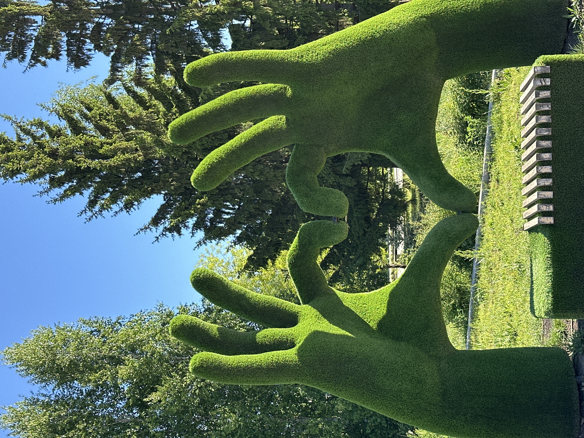 The image shows a large green topiary sculpture of two hands touching fingertips. It is situated in a garden or park, with green trees and a clear blue sky in the background. There are additional green walls with decorative wooden elements visible adjacent to the sculpture. The location is specified as the city of Unknown, Unknown.