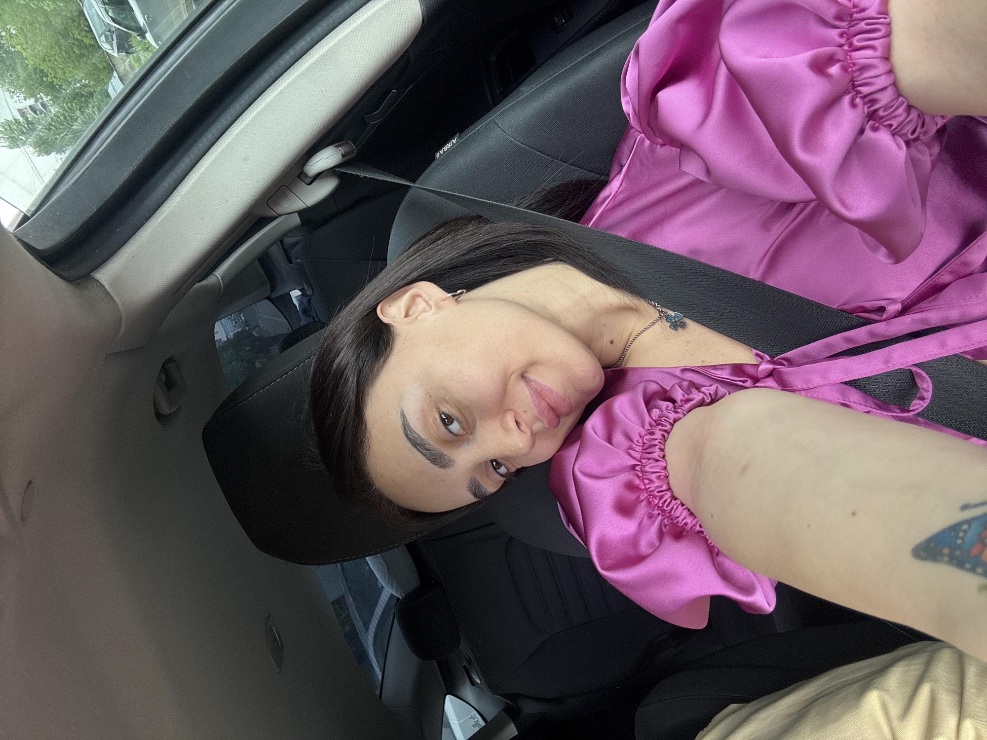 A woman is seated in the passenger seat of a car, wearing a pink satin dress with puffed sleeves and a seatbelt. She is looking at the camera with a slight smile. The car's interior is visible, including the seat, airbag label, and window. The exact location in Tbilisi, Georgia, is not discernible from the provided image.