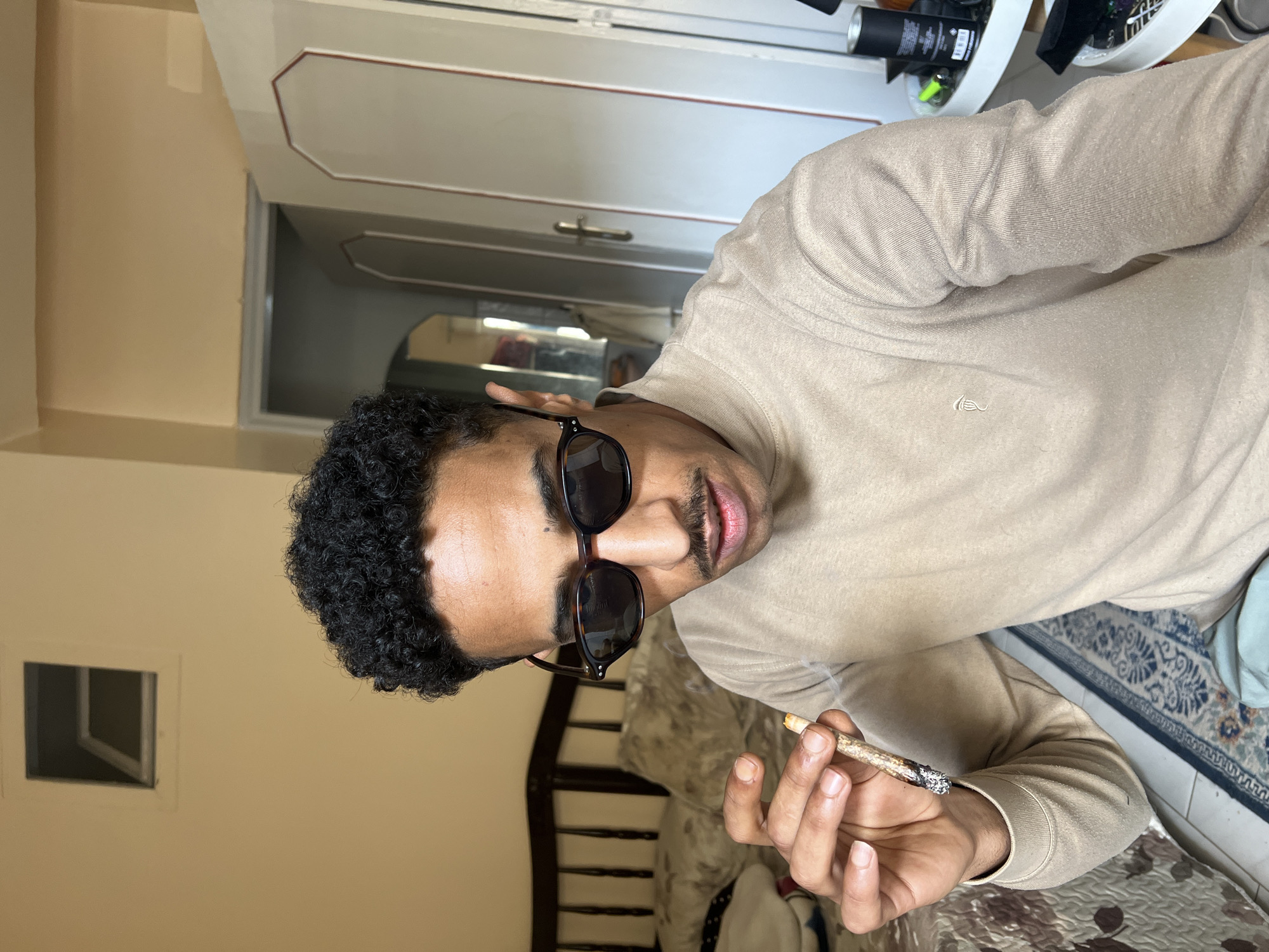 An individual is depicted indoors within a residential room in Tanger, Morocco. The subject is a male with dark, curly hair and a moustache, wearing dark tortoiseshell sunglasses and a light brown, ribbed turtleneck sweater with a small embroidered logo on the left chest. He holds a lit, hand-rolled cigarette or joint in his right hand, from which smoke is visibly emanating. The background features a dark wooden bed frame with patterned bedding on the left, and a partially open light-colored door with a brass handle on the right, revealing an interior hallway or room beyond. Walls are light beige, with a small rectangular high window opening visible in the upper left. The floor is tiled and partially covered by a patterned blue, white, and brown rug. A multi-tiered white side table holding various objects is partially visible on the far right.