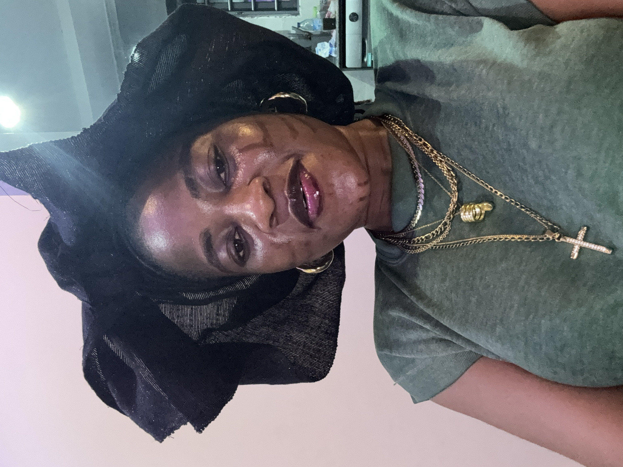 The media file is a portrait of a woman wearing a black headwrap and a green t-shirt. She is adorned with gold necklaces, one featuring a cross pendant and a ring. The woman has makeup on her lips and appears to have markings on her cheeks. She is looking towards the camera with a slight smile. The background is indistinct, but suggests an indoor setting with some light sources visible. The overall impression is one of a casual portrait taken indoors in Ajara, Nigeria. There is no visible text in the image.