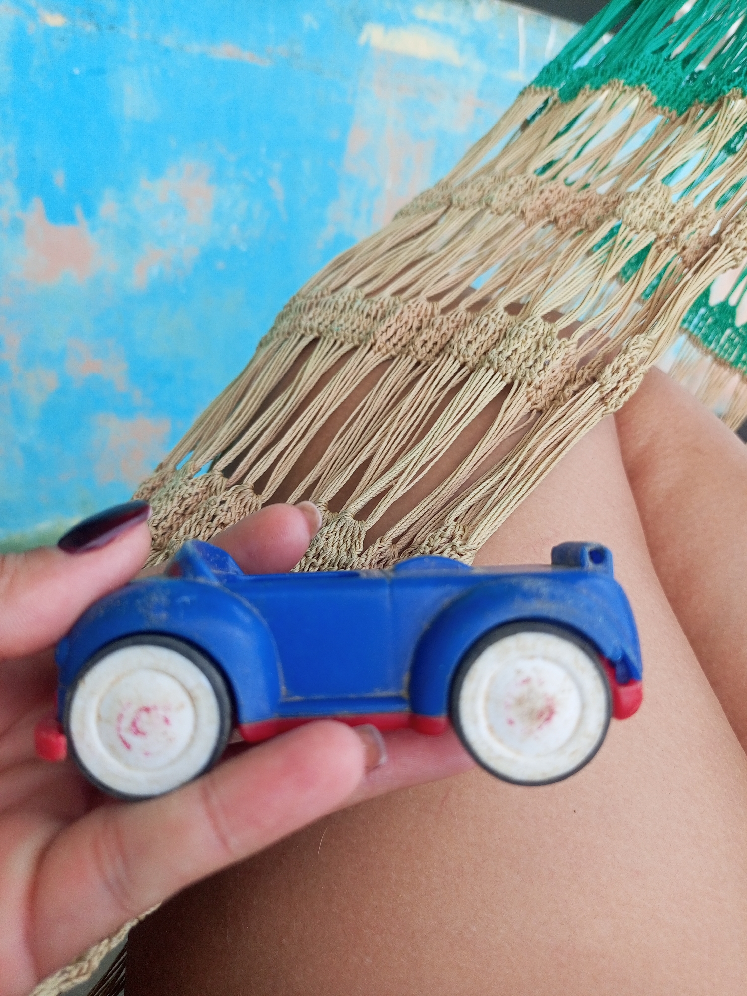 In La Tagua, Venezuela, a person's hand holds a blue toy car with white wheels and red accents. The car is positioned in the foreground, partially obscuring the hand that supports it. In the background, a hammock with a woven pattern of beige and green strands is visible, suspended above what appears to be a blue and peach-colored wall. The overall scene suggests a relaxed outdoor setting, possibly a porch or patio, given the presence of the hammock and the informal nature of the objects.