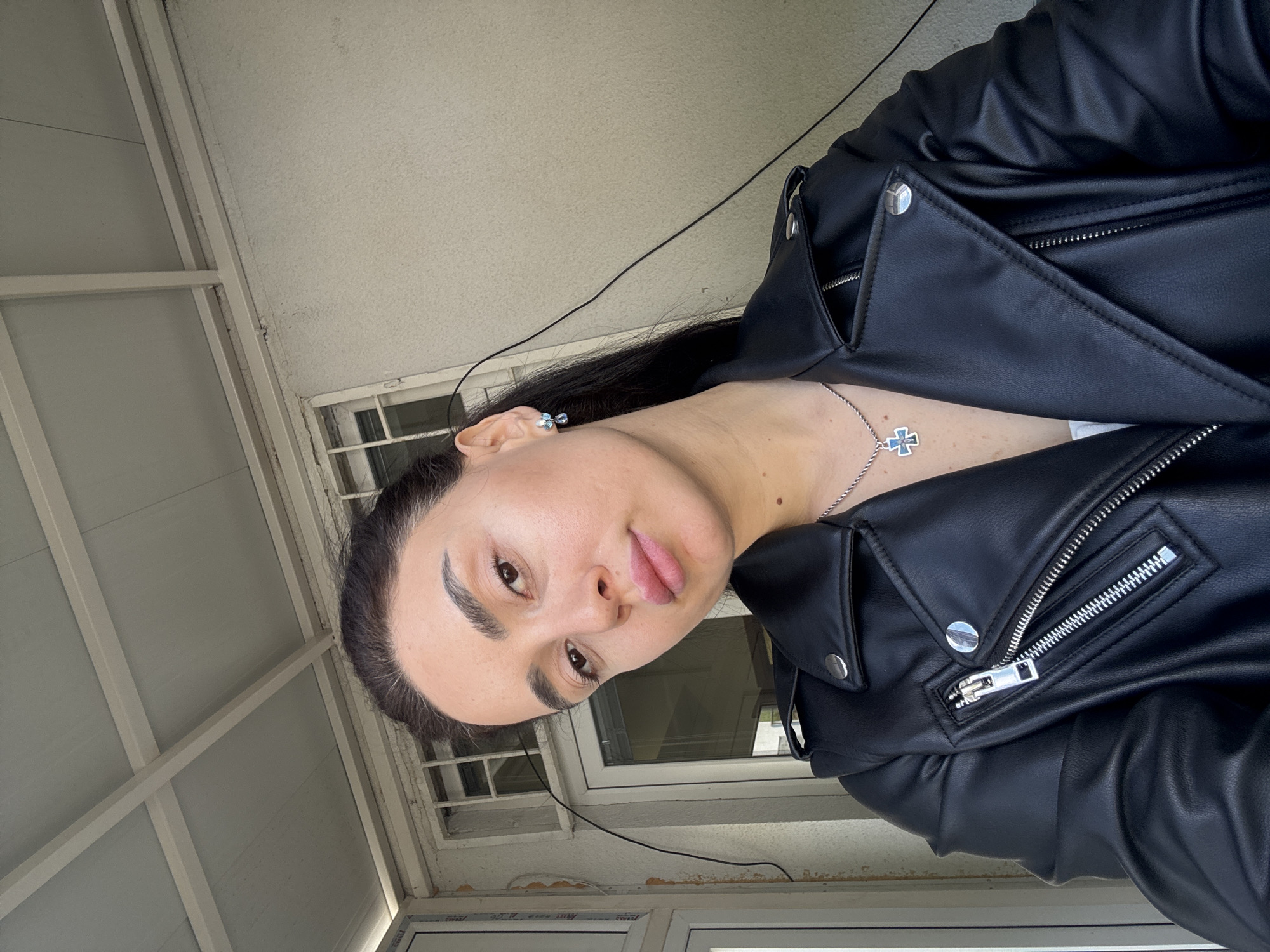 A woman in a black leather jacket stands in front of a window. The location is Tbilisi, Georgia. The woman has dark hair pulled back, and she is wearing a cross necklace and earrings. The window is bordered by a white frame and has multiple panes. Overhead, there is a white ceiling with exposed beams.