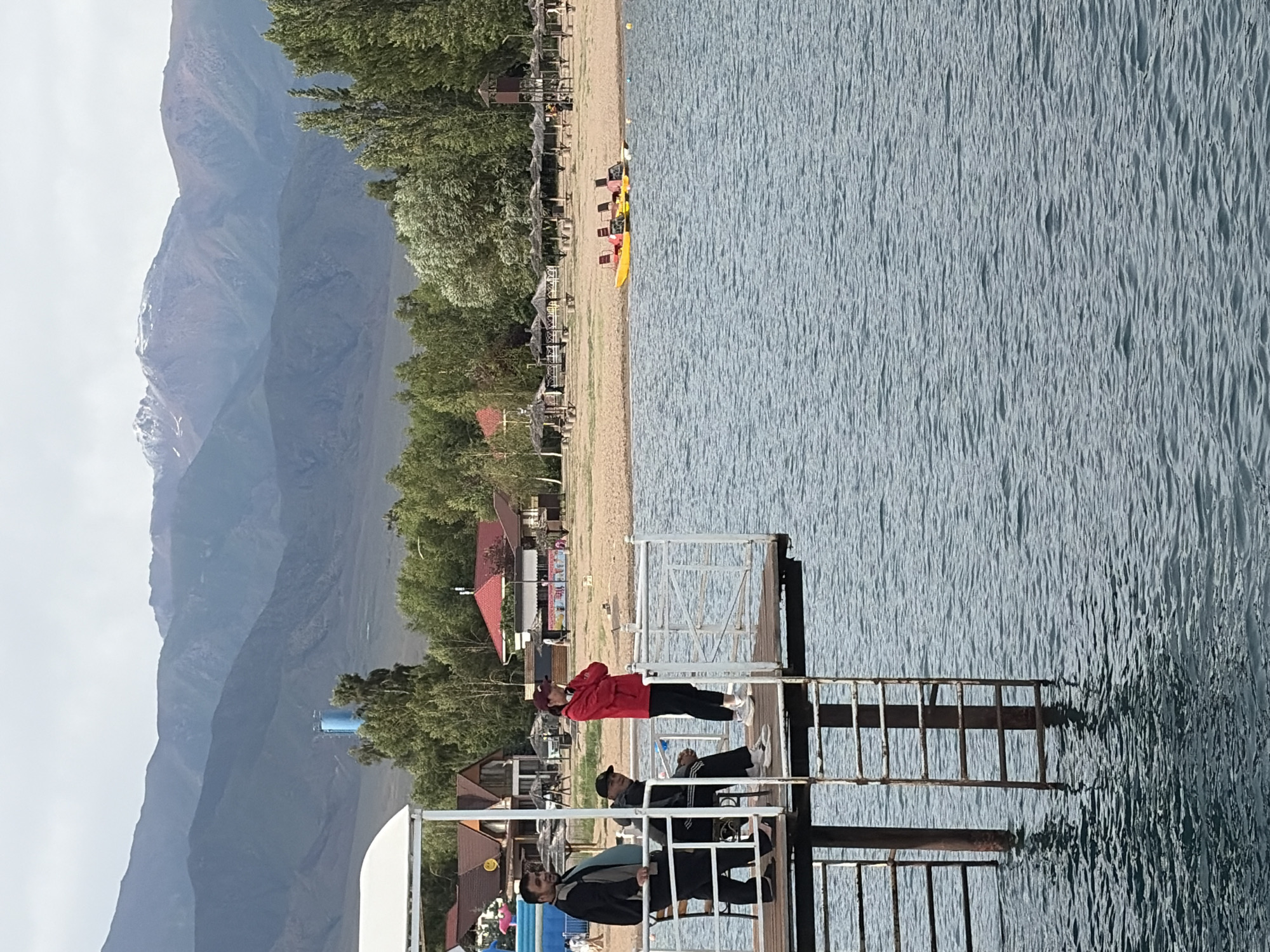 This media file depicts a picturesque scene of a lakeside resort. The primary subjects are several individuals enjoying their leisure time at the water's edge. In the foreground, a wooden pier extends into the lake, with three men visible. One man in a red jacket stands, looking towards the lake, while two others are seated, one appearing to converse with the other. Further along the shore, a sandy beach is lined with lounge chairs and umbrellas, suggesting a recreational area. Several colorful kayaks are also present on the beach, indicating water sports are available.
Behind the beach, a dense line of trees creates a natural backdrop, and beyond them, imposing mountains rise, their slopes partially covered in mist or clouds. The lake itself is a prominent feature, its surface rippling and reflecting the light, creating a textured expanse of blue. The scene appears to be set during daylight, with the sun casting shadows on the pier, suggesting it could be midday or early afternoon. The weather seems pleasant, with a clear sky visible in the far background, though the mountains suggest a potentially cooler or higher altitude environment. The overall mood is one of relaxation and outdoor enjoyment. No visible text is present in the image.