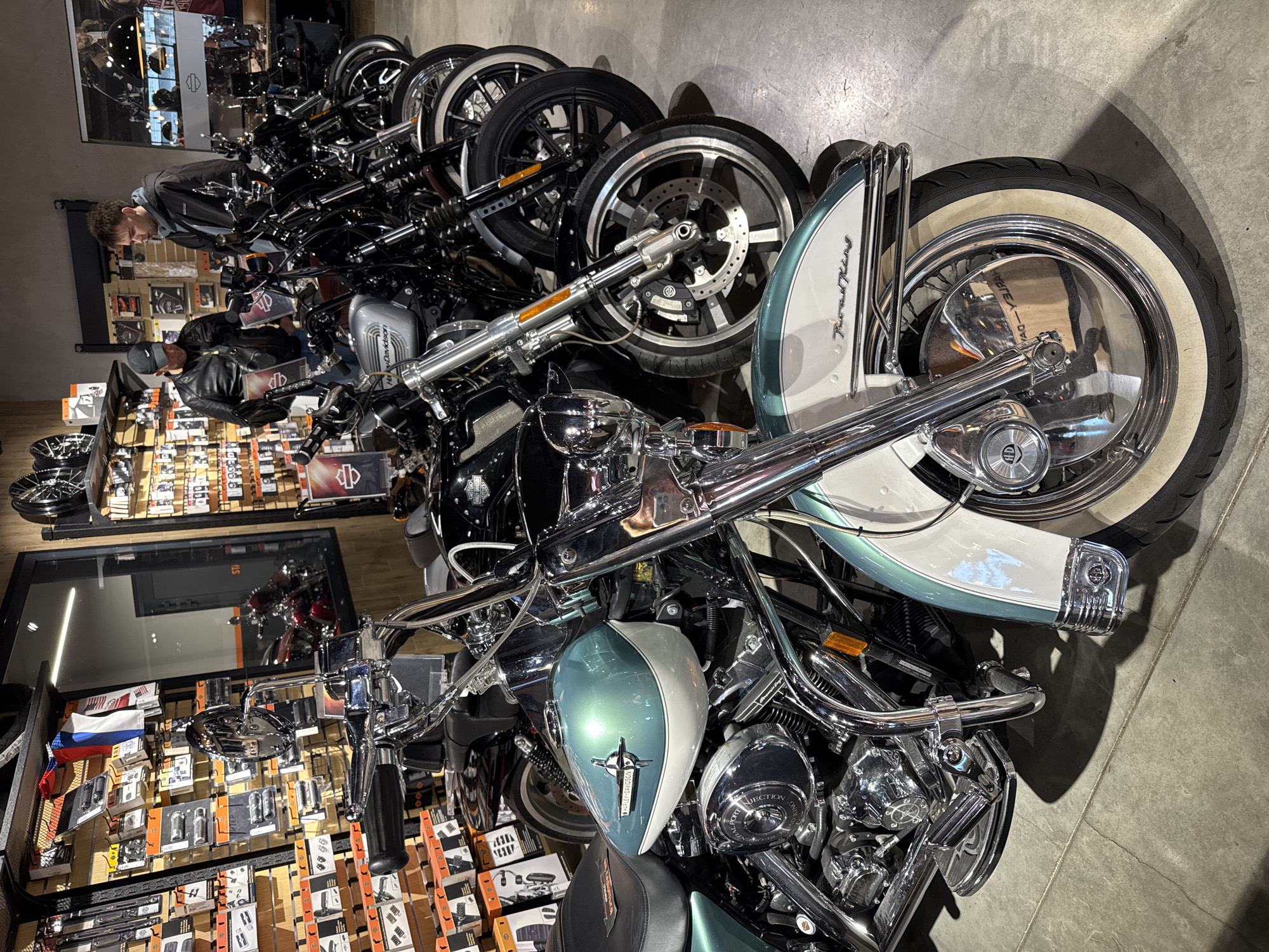 The image shows a row of Harley-Davidson motorcycles parked in what appears to be a dealership. The motorcycles vary in style and color, with chrome details prominent. One motorcycle in the foreground has a white and teal paint scheme. The location is likely within the city of Saint Petersburg, Russia, given the presence of Harley-Davidson merchandise on display. A person wearing a gray hoodie is observing the motorcycles. The environment appears to be indoors, with tiled flooring. Shelves filled with parts and accessories are displayed in the foreground on the left.