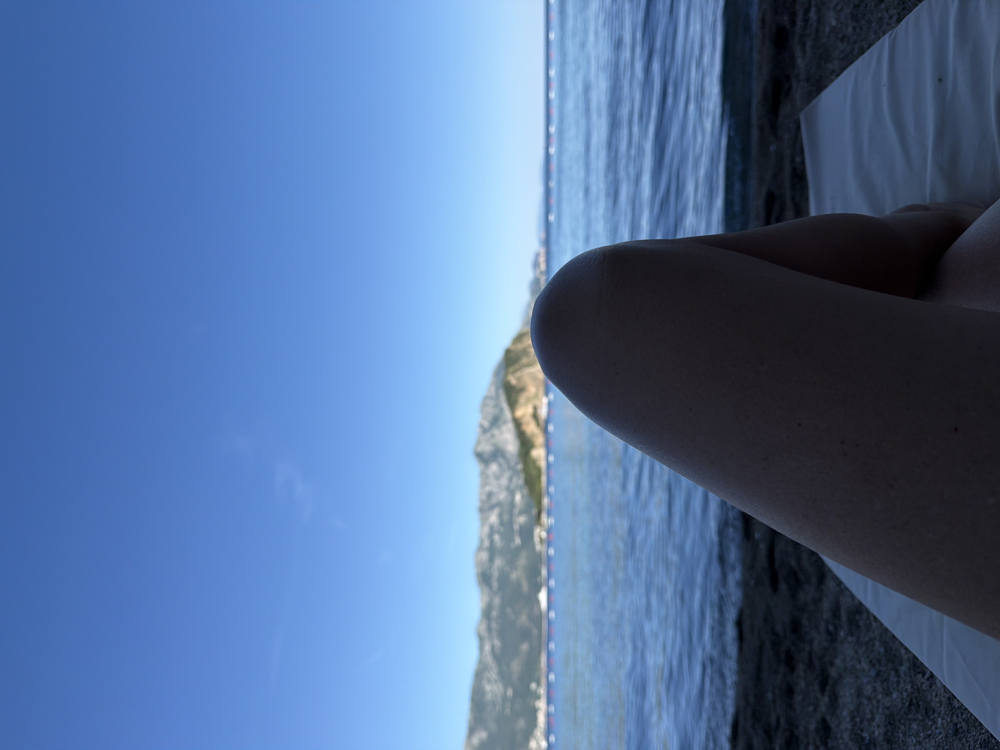 The image shows a view from a beach in Budva, Montenegro, under a clear blue sky. The scene includes the sea with a line of red and white buoys, a mountainous coastline with buildings visible at its base, and what appears to be a person's leg resting on a white beach towel. The viewpoint is at ground level, tilted to show both the sky and the beach.