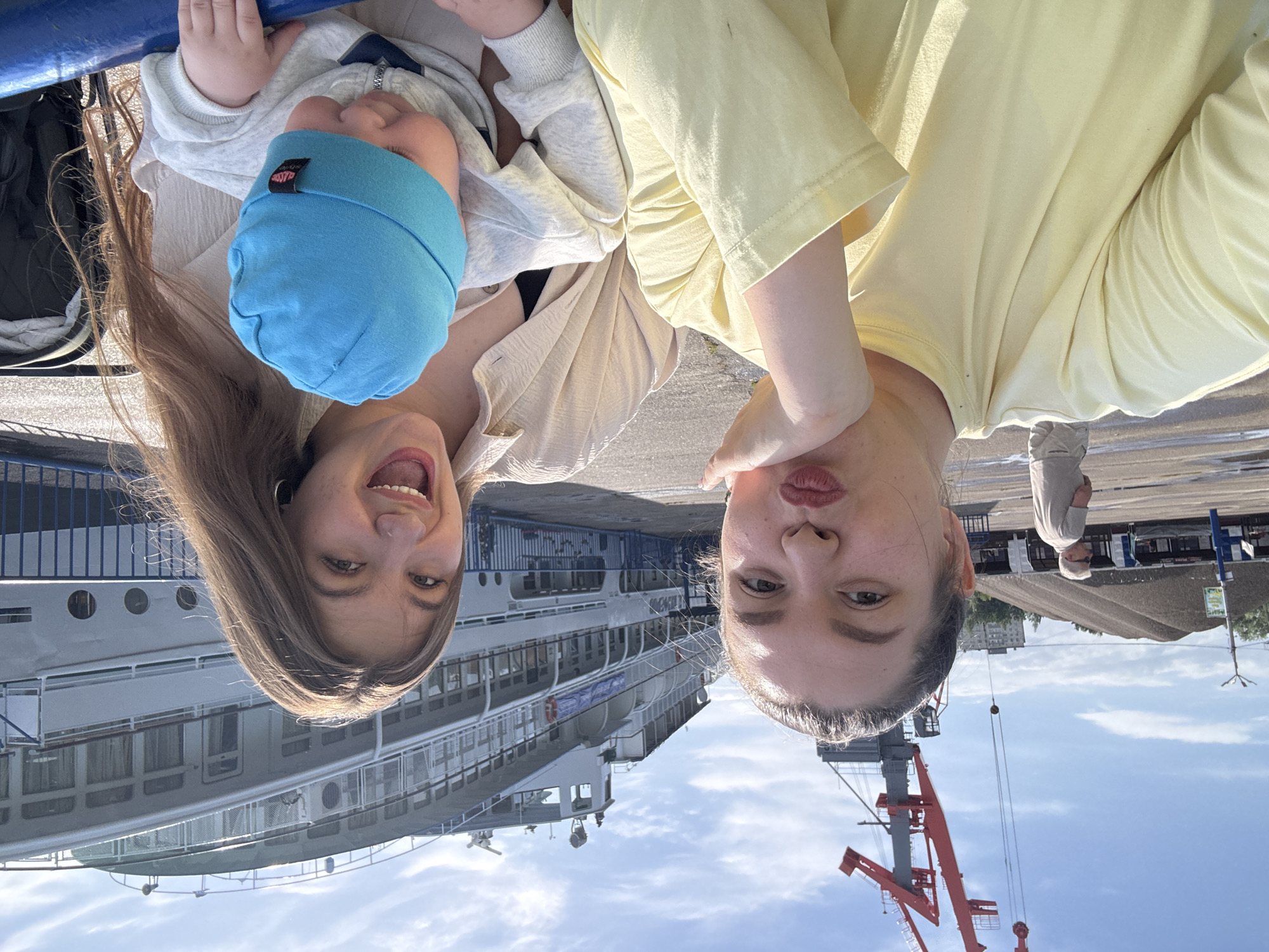 The image shows an upside-down selfie taken in Chaikovsky, Russia, with two women and a baby. The woman on the left has long brown hair and is smiling; she is holding a baby wearing a blue beanie. The woman on the right is wearing a yellow shirt and has her lips pursed. In the background, there is a ship docked along a pier, a building, a crane, and a man walking on the pier. The sky is blue with clouds.