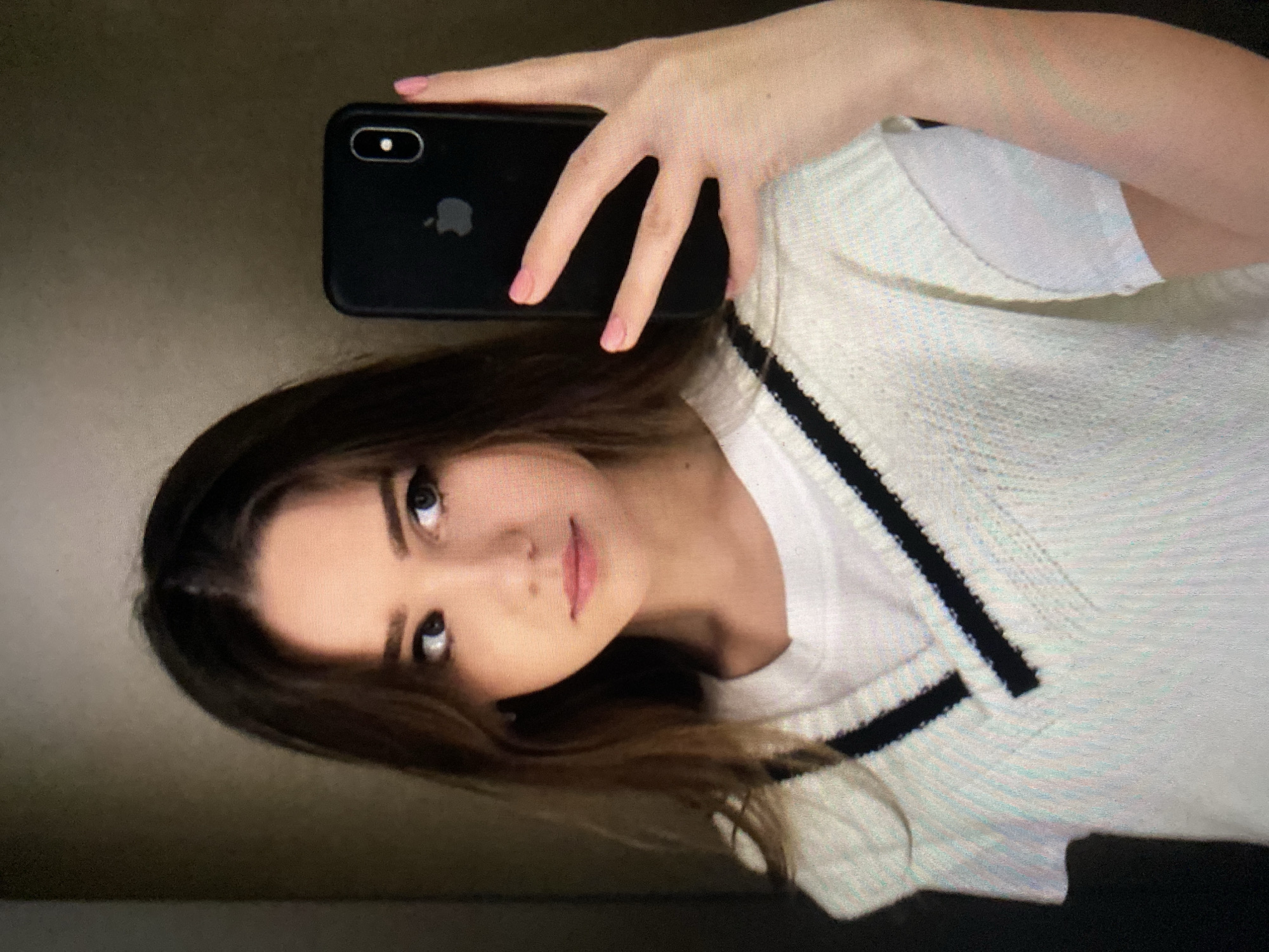 In this close-up, waist-up selfie, a young woman with brown hair and fair skin is pictured. She is looking to her right, away from the camera, with her lips slightly parted. She is wearing a white t-shirt layered under a white knitted vest with black trim. Her right hand is holding a black smartphone, with its screen facing the viewer, at chest level. The phone has a visible Apple logo. The background is a blurred, dark brown texture, suggesting an indoor setting. The lighting is soft, casting subtle shadows on her face.