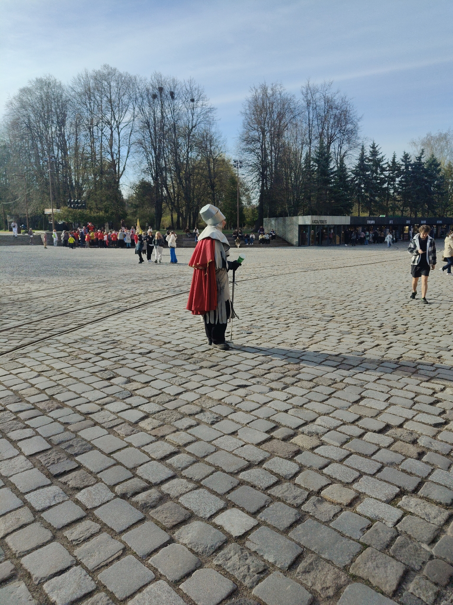 The image depicts a bright, sunny outdoor scene on a large, open plaza paved with irregular grey cobblestones. The clear blue sky overhead suggests a midday or early afternoon setting, with distinct shadows cast across the ground.
In the mid-ground, a central figure stands facing to the right, with their back partially turned towards the camera. This individual is dressed in historical or theatrical attire, featuring a light grey, possibly armored, tunic or jerkin, dark boots, and a vibrant red cape draped over their left shoulder. A distinctive light grey, almost conical helmet or coif with a subtle crest covers their head. They hold a staff or mace, topped with a green accent, in their left hand.
In the background, a line of tall, deciduous trees with bare branches suggests early spring or late autumn, interspersed with some evergreen trees. To the left, a gathering of people, some wearing red, are visible, possibly participating in an event or tour. To the right, several modern, kiosk-style buildings are present. On one building, the text "KACCA/TICKETS" is clearly visible, indicating a cashier or ticket booth. Another sign reads "СУВЕНИРЫ" (Souvenirs), with additional partially visible signs for "GIFTS" and "COFFEE". People are seen walking around these buildings and across the cobblestone plaza, including one individual in a plaid shirt and shorts. The scene appears to be a public square, possibly during a festival or public gathering, with the costumed figure adding a unique point of interest.
