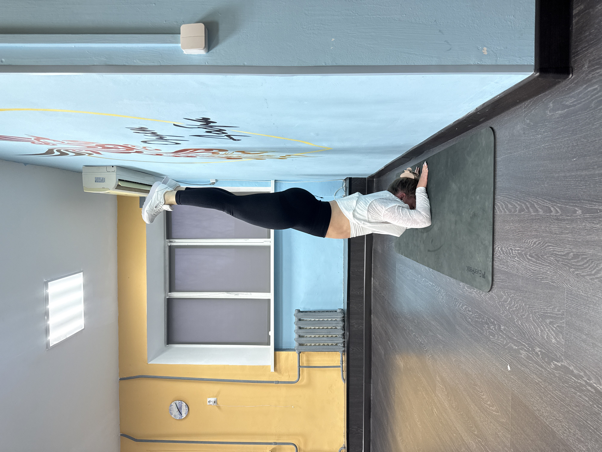 A woman is performing a forearm plank on a gray yoga mat in a room with a unique color scheme. The upper part of the wall is light blue, decorated with painted designs, and the lower part is yellow. The floor is gray wood laminate. The woman is wearing black leggings, a white top, and white sneakers. There is a window, radiator, and clock visible on the wall. An air conditioning unit is mounted high on the wall. The image was taken in Chaikovsky, Russia.