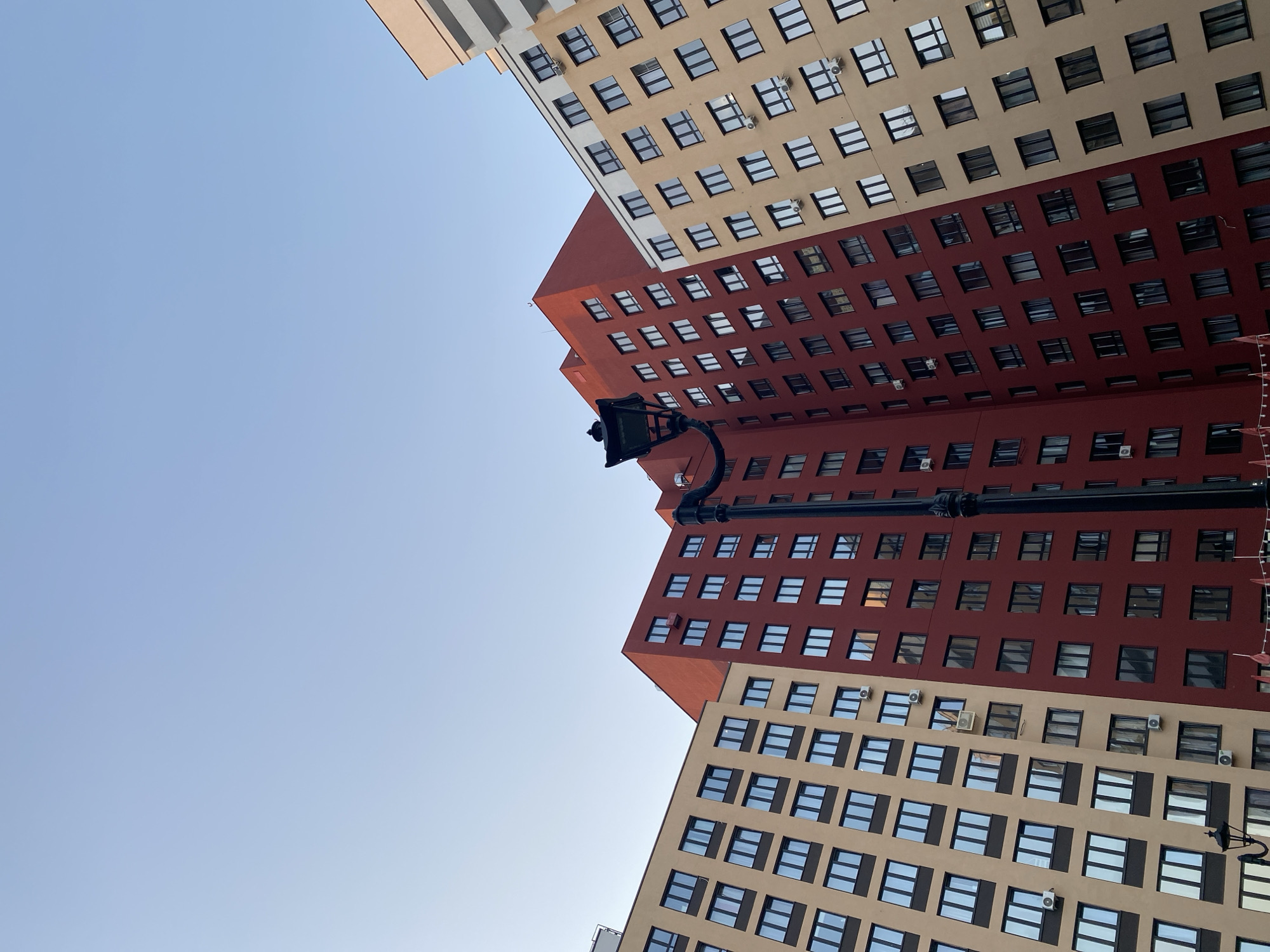 The image captures an upward view of three modern buildings in Yekaterinburg, Russia. These buildings have repeating window patterns with red-brown and beige sections on their facades. A black street light post is centrally positioned in front of the buildings. The sky above is a clear pale blue.