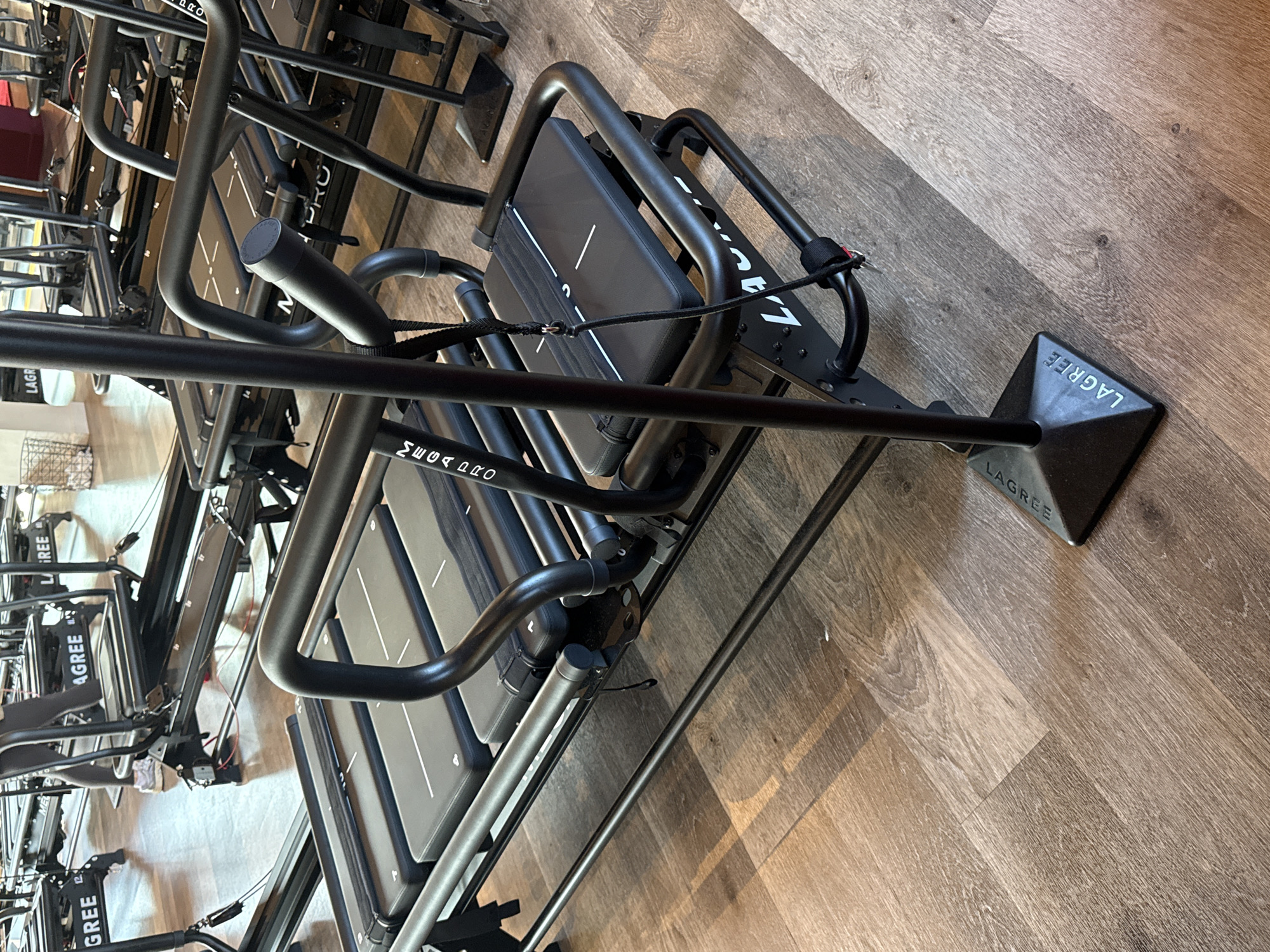 The image displays an indoor fitness studio, identified as a Lagree Fitness center, likely situated in London, United Kingdom. The foreground features a black Lagree Megaformer exercise machine, characterized by its black metal tubular frame, multiple padded platforms, and curved handlebars. A prominent vertical pole with a triangular base, embossed with "LAGREE," extends from the machine's front and rests on a light brown wood-effect floor. A black fabric strap with a metallic clip hangs from one of the handlebars. In the mid-ground, several additional identical or similar black Megaformer machines are arranged in rows, displaying "LAGREE" and "MEGAPRO" branding. A partial view of an individual's lower legs and feet, clad in dark leggings and white athletic shoes, is visible standing between two machines. The environment consists of light-colored walls and appears brightly lit. A white wire basket is partially visible in the distant background. No direct actions are depicted.