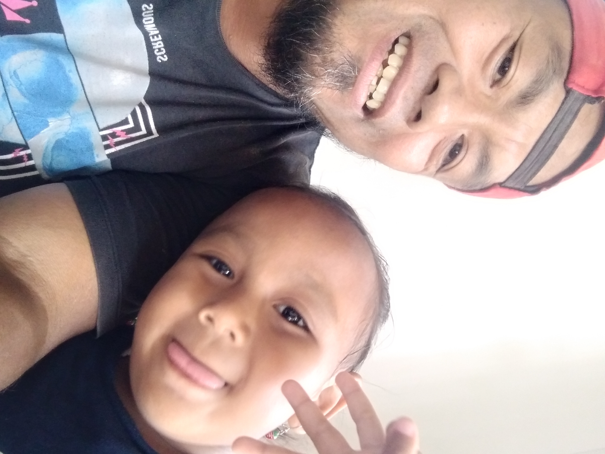 This is a selfie taken by a man wearing a red baseball cap and a black t-shirt. He is smiling widely, showing his teeth, and has a goatee with graying hairs. To his right, a young child rests their head on his shoulder. The child is also smiling and has their tongue sticking out slightly. The background is plain and light-colored, suggesting an indoor setting. The man's t-shirt has a graphic design on it. The image appears to be taken in Pandak, Indonesia.