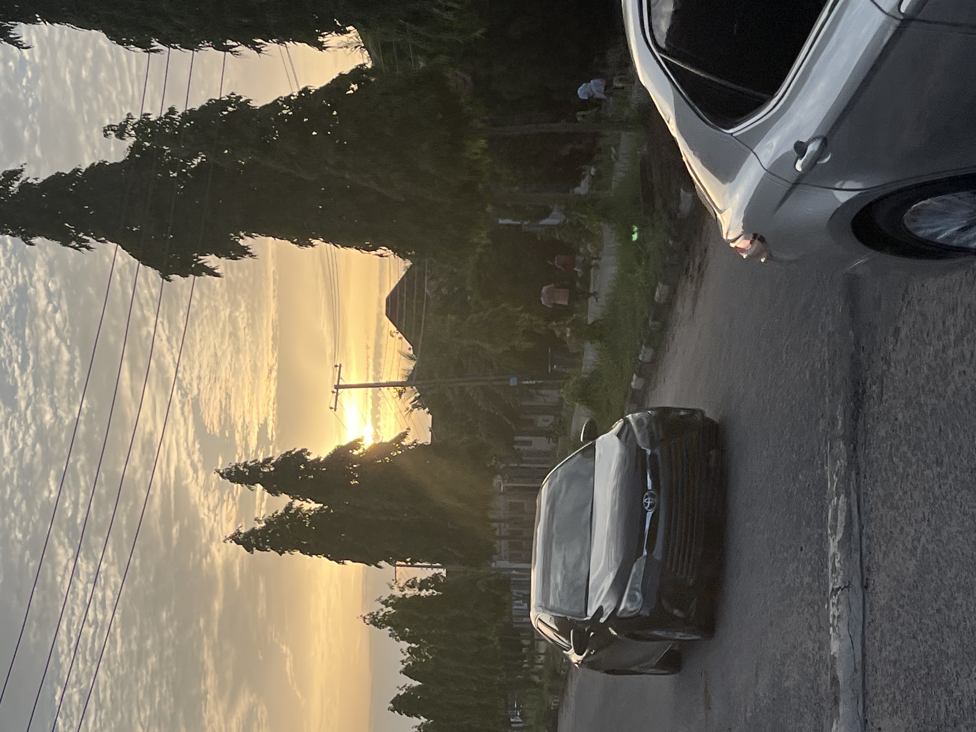The image captures a street scene in Abuja, Nigeria, at what appears to be sunset. The sky is a mix of orange and grey clouds. Two cars are visible on the street: a silver car is partially in view to the right and a black car in the middle of the street. Trees are on either side of the road, and power lines are in the upper left of the frame. A building is visible in the background between the trees. People are walking on the sidewalk in the distance. The road is paved and has a white line marking the edge.