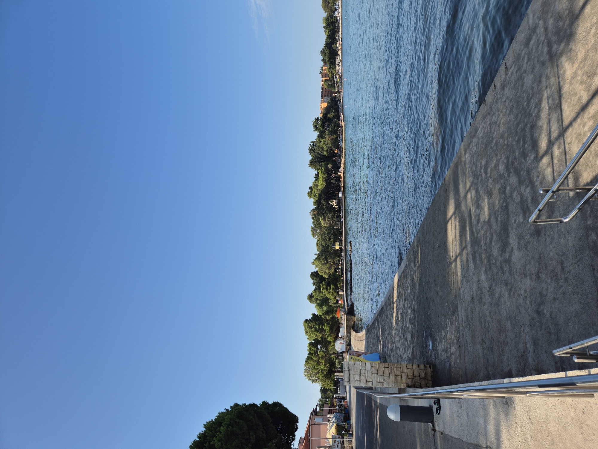 The image depicts a coastal scene in Monterol, Croatia, under a clear blue sky. In the foreground, a concrete promenade runs along the edge of a body of water, with metal railings and stairs visible. The promenade is bordered by a low stone wall on the left, separating it from a paved road and sidewalk. A dark bollard with a white top is present on the sidewalk.
Further along the promenade, a person in a blue garment is walking. The background features a line of trees and vegetation, with buildings peeking through the foliage. Some buildings appear to be houses or small establishments along the coast, and a larger, orange-hued building is visible in the distance. The water is calm with gentle ripples. The overall impression is a serene seaside environment, likely a public walkway or recreational area.