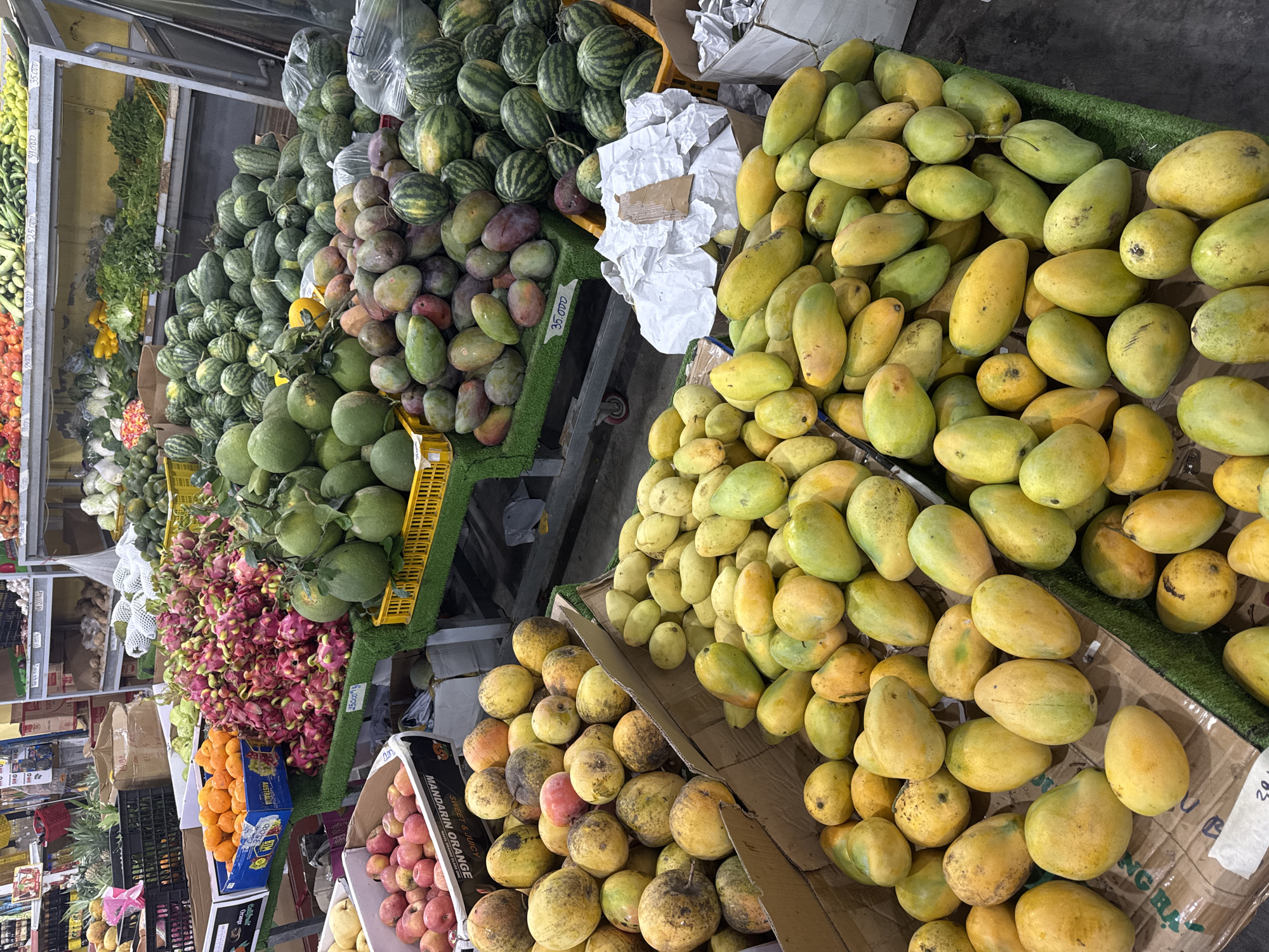 This is a high-angle, full shot of a vibrant fruit stall in Vietnam. The scene is overflowing with a diverse array of fresh produce, primarily displayed in open boxes and on shelves. Prominently featured are large piles of yellow and green mangoes, filling two large cardboard boxes. To the left, a substantial quantity of watermelons is neatly arranged, some with yellow stickers. Below the watermelons, a colorful display of red and pink dragon fruit draws the eye, nestled beside a crate of bright orange tangerines. Further left, rows of green cucumbers and other vegetables are visible on shelves, alongside what appear to be bags of produce and some packaged goods. A yellow plastic basket is also visible, filled with green fruits. The overall impression is of a bustling market, abundant with fresh, seasonal offerings, likely during daylight hours given the bright lighting. The visible text includes brand names like "Callfruit," "Orange," "MANDARIN ORANGE," and prices such as "35.000" and "25.000", indicating this is a commercial setting. The sheer volume and variety of fruits and vegetables suggest a well-stocked market stall catering to a local clientele.