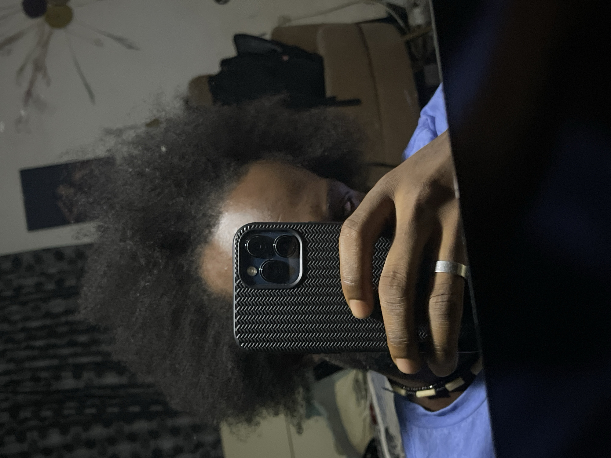 A person with dark skin and afro-textured hair is taking a selfie in a mirror with a black smartphone. The person is wearing a light blue shirt and a ring on one finger. The smartphone has a dark case with a textured pattern. A beaded necklace is visible. Background details include a ceiling fixture, a dark framed object on the wall, and furniture. The location of Kuchingoro, Nigeria is not evident from the image.