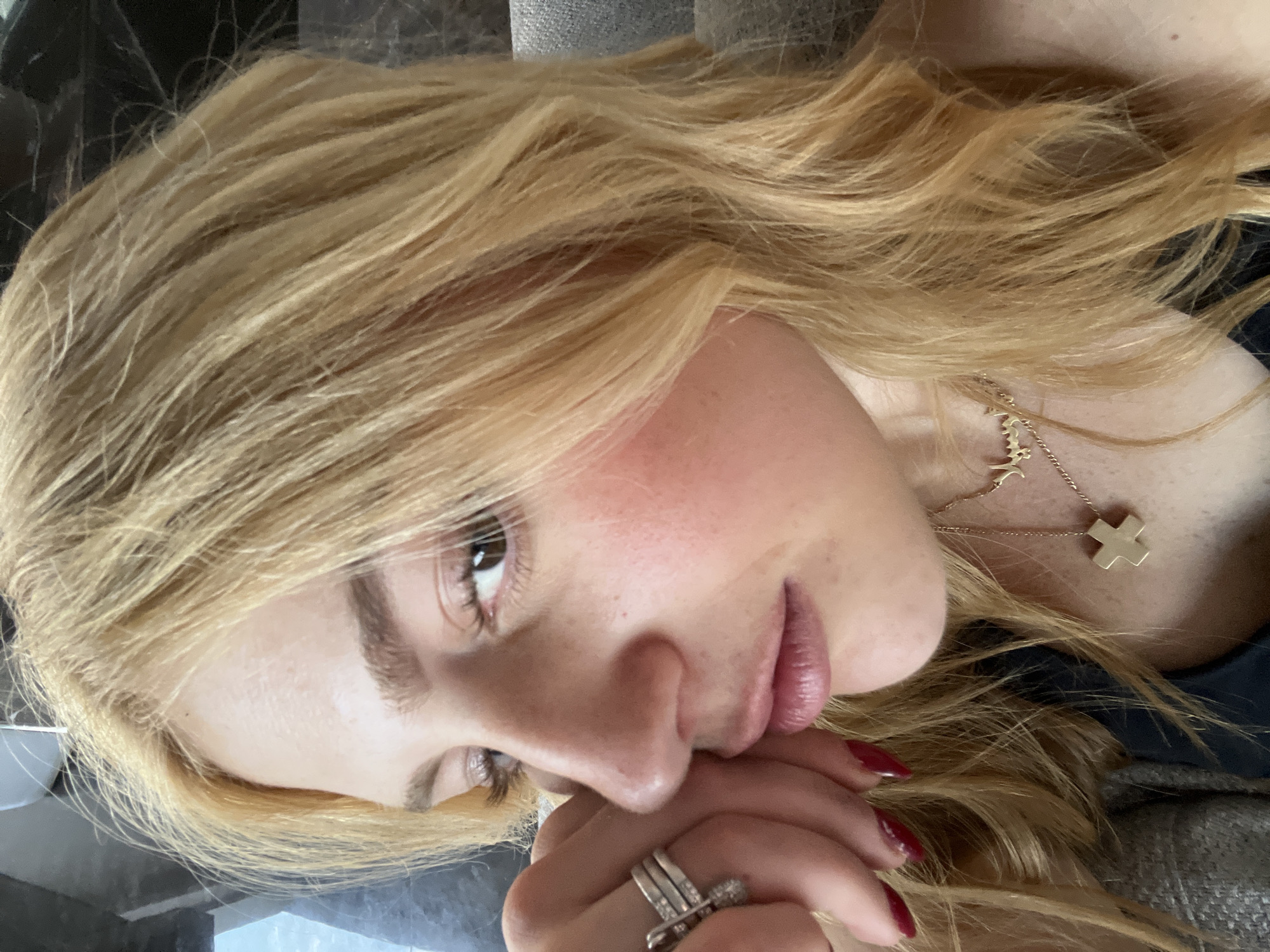 A close-up portrait features a female subject with fair skin, light freckles, and long, wavy, golden blonde hair falling over her shoulders. Her brown eyes, enhanced with elongated eyelashes and a light, shimmery eyeshadow, gaze directly forward. Her full lips are colored with a reddish-pink product. Her left hand, with dark red polished fingernails and multiple silver-toned rings on her fingers (including one multi-band ring), rests against her left cheek and jawline. She wears a dark garment at the neckline and two gold-toned necklaces: one with a small cross pendant and another with a script-style pendant. The background is out of focus, revealing a dark, textured surface behind her head and a light grey, woven fabric, consistent with upholstery, to her right, indicating an indoor setting. This scene is located in Santiago Yancuitlalpan, Mexico.