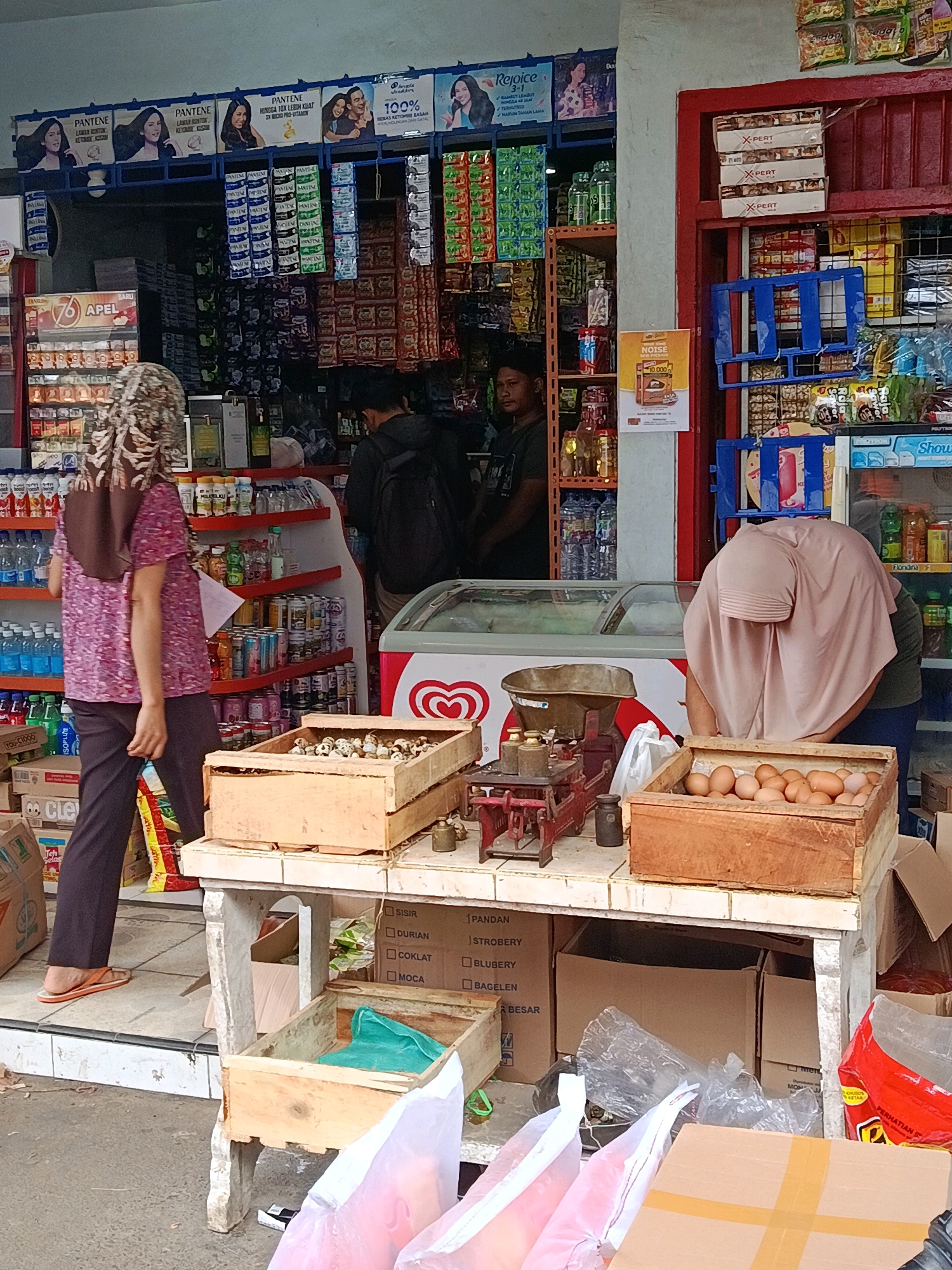 A street-level shot captures a roadside stall in Sidorenggo, Indonesia, likely a small general store or warung, showcasing an array of goods. A woman, wearing a patterned purple shirt, brown headscarf, and dark pants, is walking away from the camera towards the left, her orange flip-flops visible. She carries a brown bag on her shoulder. Another person, partially obscured by a large pink hijab, is bent over a wooden crate filled with eggs, her face hidden from view. The stall displays various items, including bottled drinks, packaged snacks, and a prominent red and white ice cream freezer. A vintage scale sits on a table near the egg crate, adorned with weights. The background reveals shelves packed with merchandise and advertising posters. The ground is paved with grey concrete and tiling. The scene appears to be set during the day, with natural light illuminating the area.