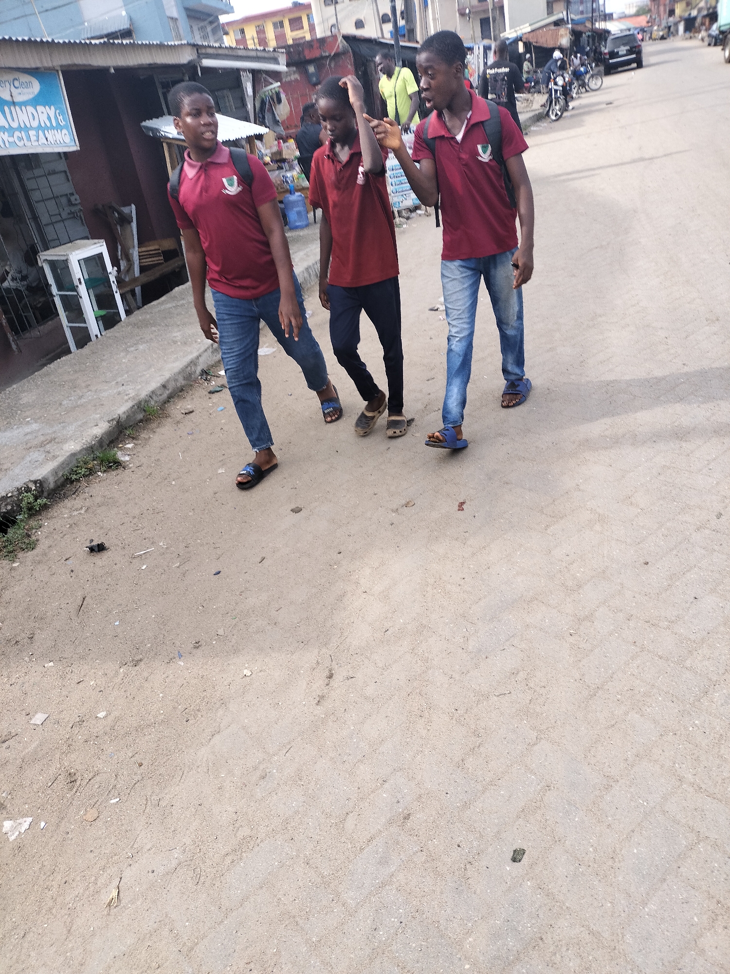 The image depicts three young males walking along a sunlit street in Alaiyabiagba, Nigeria. All three subjects, likely teenagers or pre-teens, are wearing similar maroon polo shirts with a crest on the left chest, suggesting a school uniform. Two wear blue jeans and carry backpacks, while the third wears dark trousers. They are all shod in open-toed sandals or slides.
The boys are walking from left to right in the foreground, on a dusty, unpaved road that transitions into a more structured, possibly paved surface further down. The boy on the far right has his right hand near his head and appears to be looking towards his left, possibly in conversation or reaction to his companions. The other two boys look forward or slightly to their left, engaged in the casual stroll.
The background shows a busy urban or semi-urban street lined with commercial buildings. On the left, a blue sign is partially visible, clearly displaying "LAUNDRY & DRY-CLEANING." Other background elements include market-like stalls, various people, motorcycles, and a black SUV in the distance. A blue plastic water dispenser is visible on the sidewalk. The bright illumination and distinct shadows indicate clear, sunny weather, likely midday or early afternoon. The overall scene portrays a slice of everyday life in a vibrant Nigerian street setting.