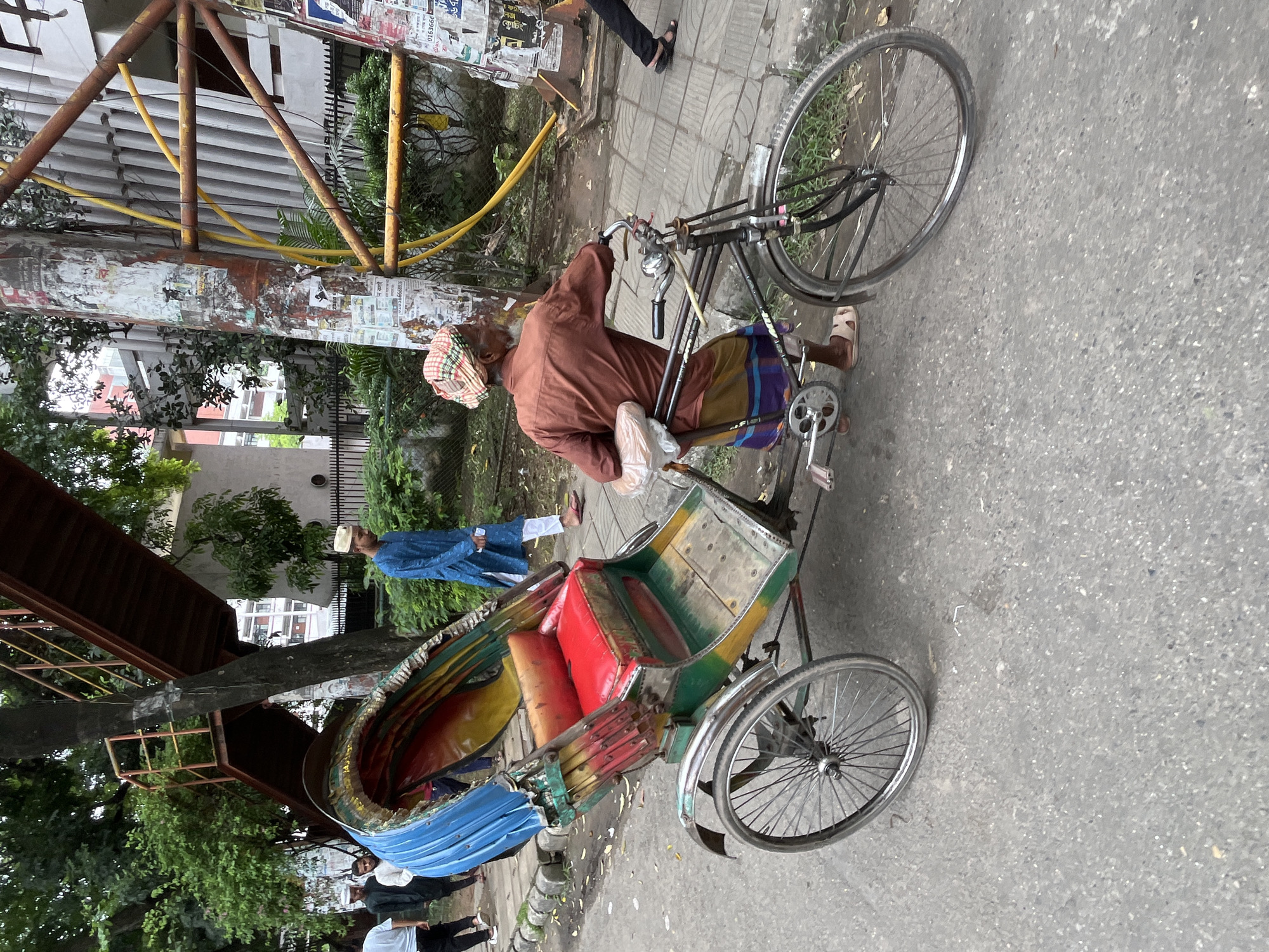The image shows a street scene in Dhaka, Bangladesh. In the foreground, a man is pushing a cycle rickshaw. The rickshaw is colorful, with a red seat and a blue and yellow canopy. The man is wearing a brown shirt and a patterned headscarf. Behind the rickshaw, other people are walking on the sidewalk. In the background, there is a metal staircase and trees. The overall scene depicts a busy urban environment.