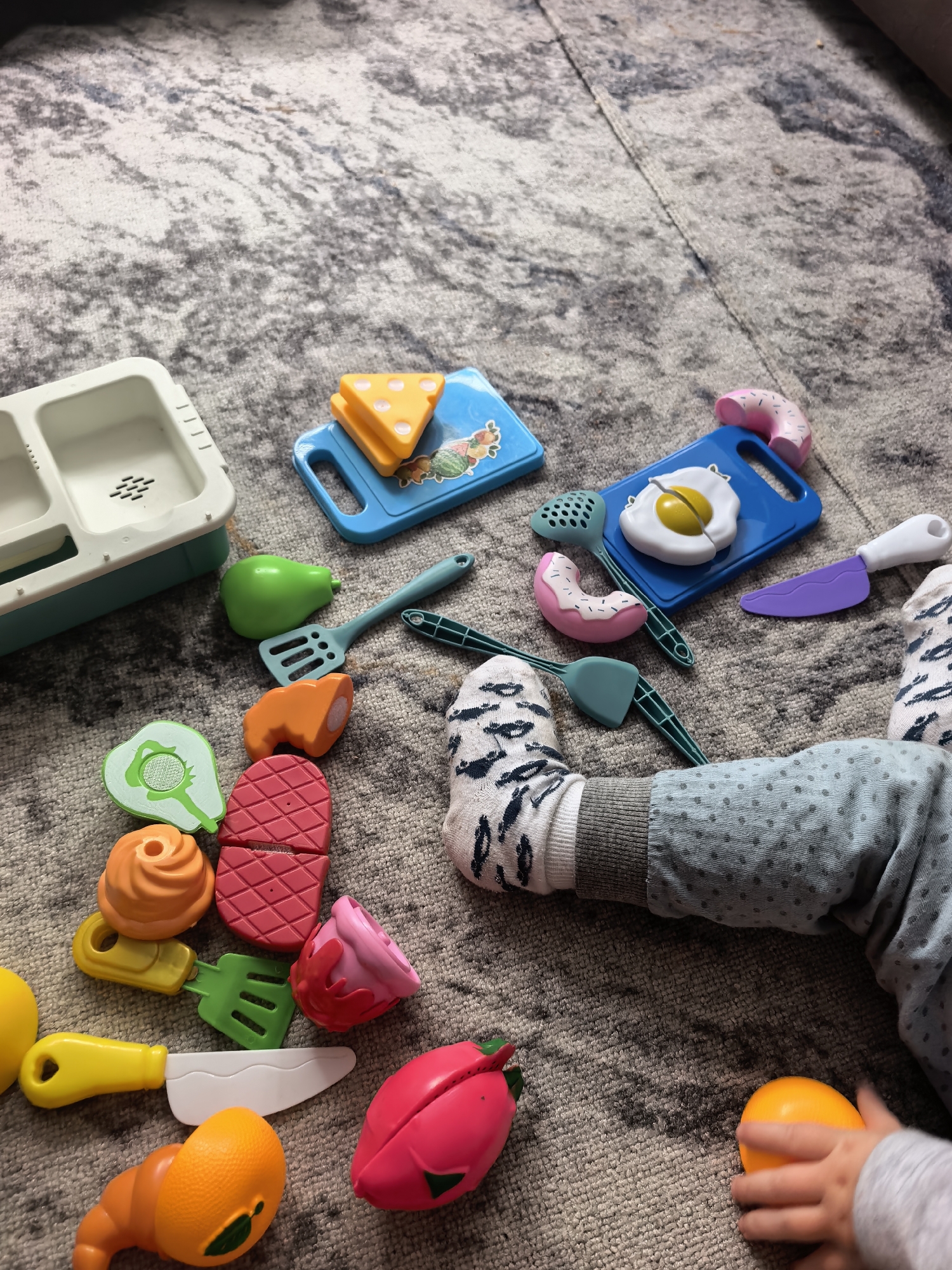 The image shows a collection of toy food items and kitchen utensils scattered on a grey carpet. A child's legs, wearing patterned socks and grey polka-dot pants, are visible in the foreground. Among the toys are a toy sink, cutting boards, a toy egg, donuts, a pear, steak, avocado, carrots, a strawberry, a croissant, and a yellow knife. The scene depicts a child playing with their toys on the floor. There is no information about the location in the city of Sterlitamak, Russia.