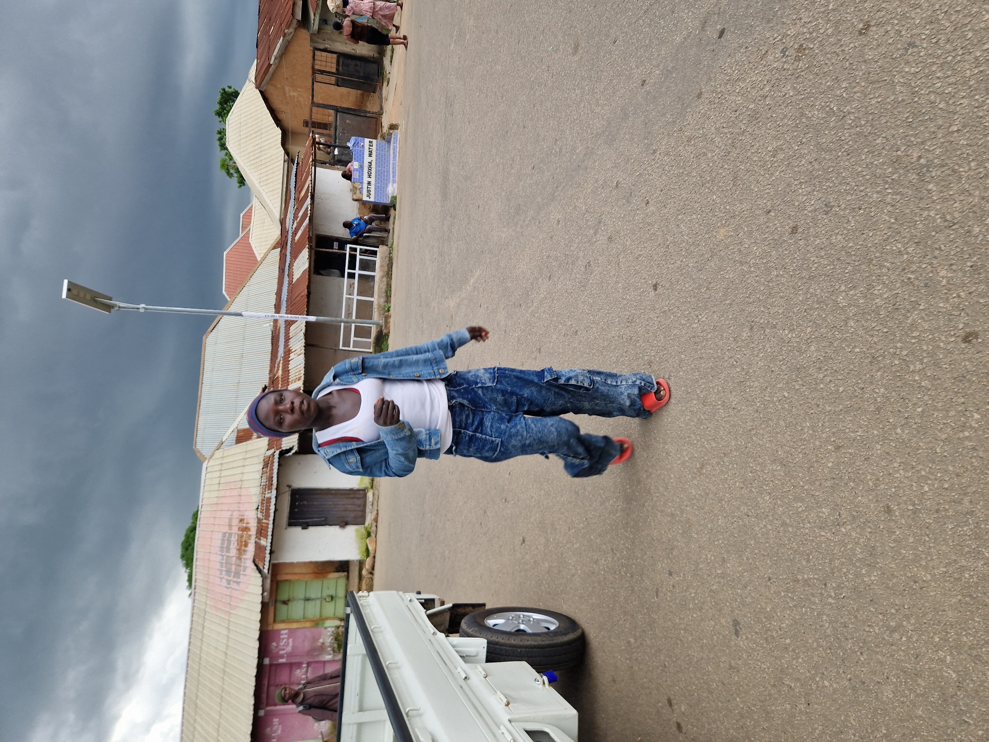 A young Black woman wearing a blue denim jacket, a white tank top, baggy blue jeans, and red flip-flops walks across a paved street. She is looking forward, slightly down. Her left hand is by her side, and her right hand is extended slightly in front of her. Behind her, a white flatbed truck is parked on the left side of the road. In the background, there are several low-rise buildings with corrugated metal roofs, some appearing weathered. A street light is visible to the left of the woman. The sky is overcast with dark clouds. A sign reading "JUSTIN HOXHA, WATER" is visible on a blue tiled structure. Other people are present in the background, including one sitting on a bench and two walking on the right side of the street carrying items on their heads. The overall setting appears to be a commercial or residential area in Kabong, Nigeria.
