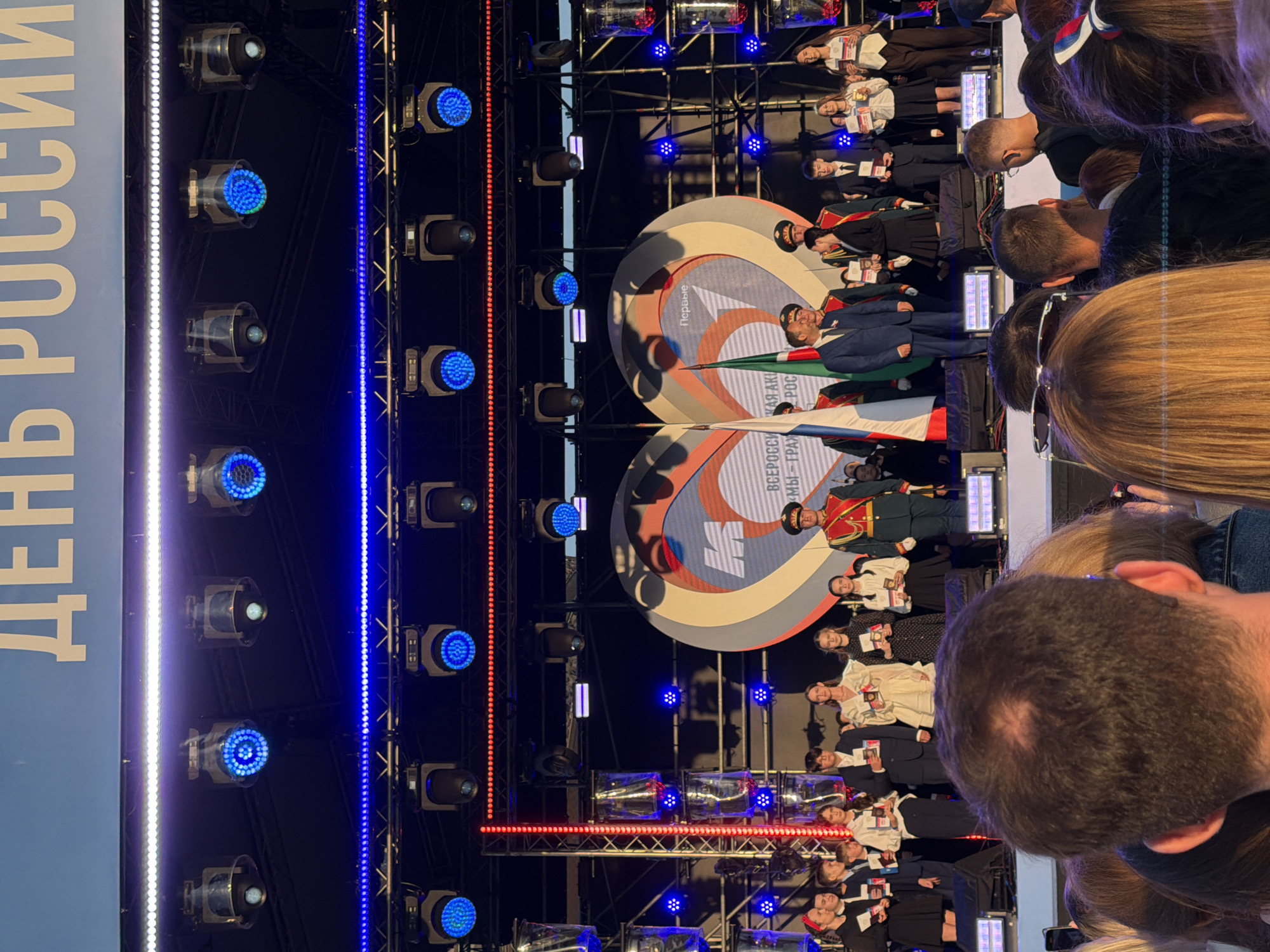 An outdoor evening ceremony is taking place on a stage. The backdrop features a large, curved screen with a logo resembling an 'M' within a heart shape, and text in Russian. A line of people, including children and adults in formal attire and military uniforms, stand on the stage, receiving or holding documents. Several flags are displayed. Above the stage, a truss system is equipped with numerous blue and white stage lights. In the foreground, the backs of several spectators' heads are visible, suggesting they are part of an audience watching the event. The overall atmosphere appears to be celebratory.