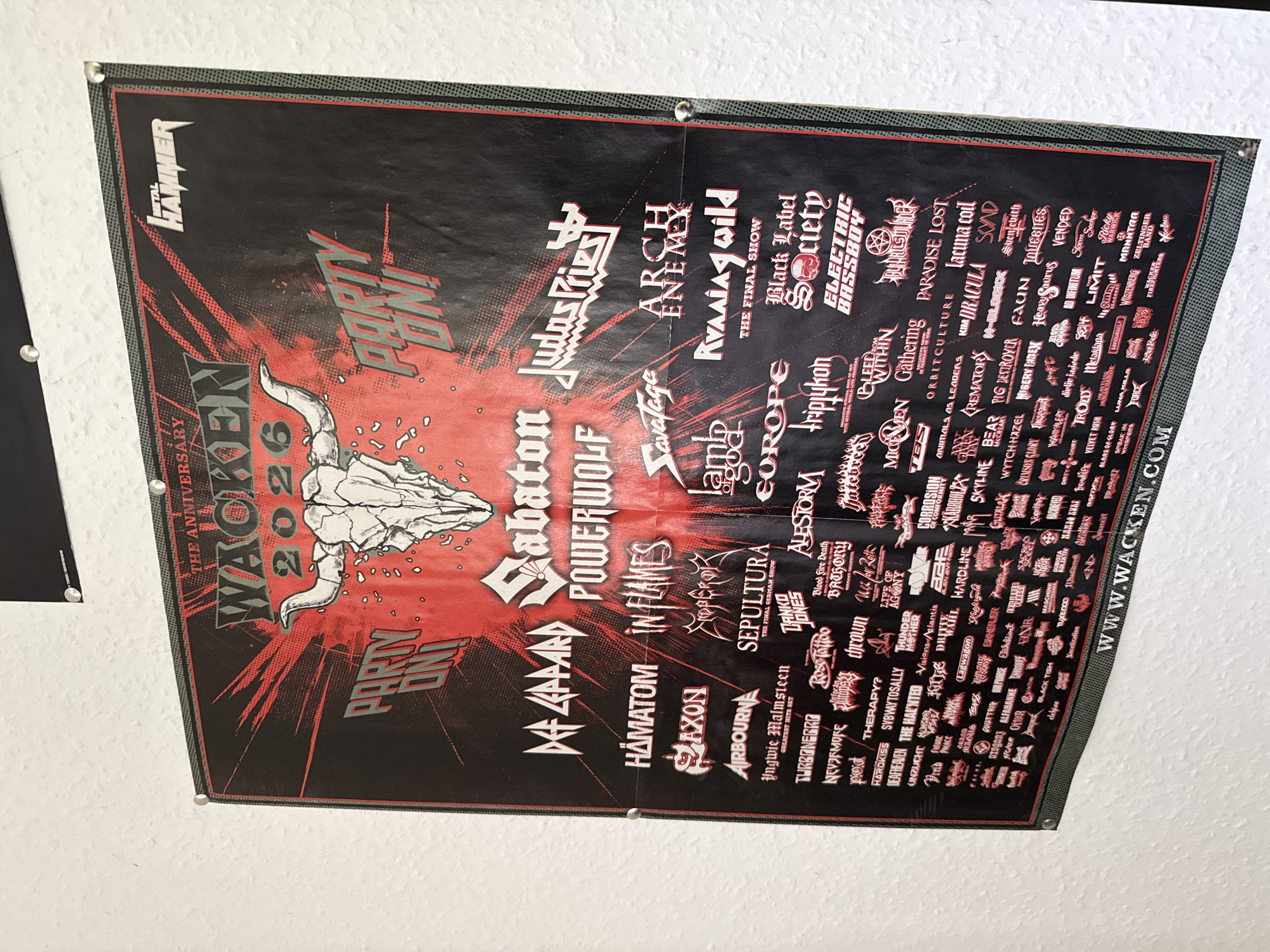 The image shows a large Wacken Open Air 2026 festival poster hanging on a textured, light-colored wall. The poster is predominantly black and red with white and grey text and graphics. At the top, it reads "THE ANNIVERSARY" and "WACKEN 2026" with a stylized bull skull graphic. The "METAL HAMMER" logo is present in the upper right corner. Prominently displayed bands include Def Leppard, Sabaton, Powerwolf, and Judas Priest. Below these headliners are numerous other band names listed in a dense grid format. The bottom of the poster features the website "WWW.WACKEN.COM". The poster is attached to the wall with small silver thumbtacks. There are no people or any other objects visible in the immediate scene besides the poster and the wall. The environment appears to be indoors, likely a room with minimal natural light. The city of Bad Sachsa, Germany is not depicted or mentioned in the visual content of the image.