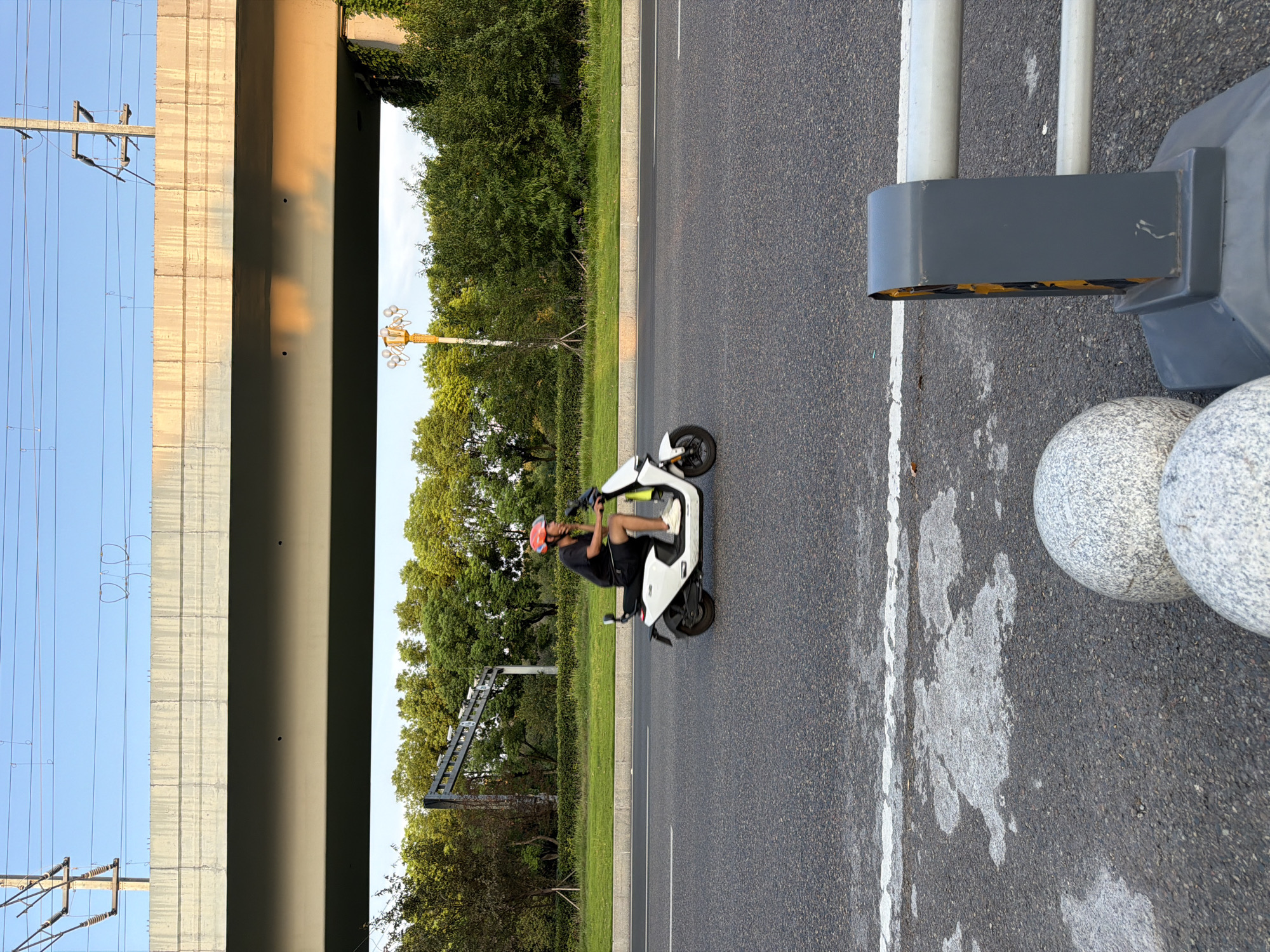 A person wearing a black t-shirt, black shorts, and a red helmet is riding a white electric scooter on a paved road. The scooter has a black seat and a white body. The rider's left hand is on the handlebars, and their right arm is resting on their lap. The scooter is in motion, indicated by the blur of the wheels.
The road is asphalt and has a white line painted on it, dividing lanes. The environment around the road consists of a green lawn and a dense line of trees. In the background, there is a large concrete overpass supported by columns, with power lines running overhead. The sky is clear and blue, with the warm glow of the setting sun visible on the underside of the overpass.
In the foreground, on the right side of the image, a gray traffic barrier with yellow accents is visible, along with two large, spherical stone bollards. The scene appears to be set in Jiangjin, China, judging by the context provided. The lighting suggests it is late afternoon or early evening.