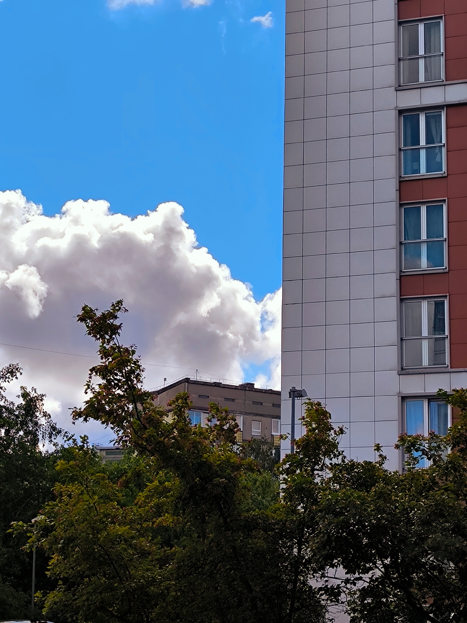 This image captures a juxtaposition of urban architecture and nature under a bright blue sky with large, fluffy white clouds. On the right, a modern building with a facade of light gray square tiles and red accents dominates the frame. Several windows with white frames and reflective glass are visible on this structure. To the left of this building, a portion of an older, less imposing building is visible, appearing to be a Soviet-era block with windows. In the foreground, dense green trees with scattered red leaves suggest the season is late summer or early autumn. A streetlamp is partially visible amidst the foliage. The scene appears to be set in an urban environment, possibly a residential area, in Riga, Latvia, given the architectural style and the characteristic large cumulus clouds often seen in the Baltic region. There are no people or discernible actions in the image.
