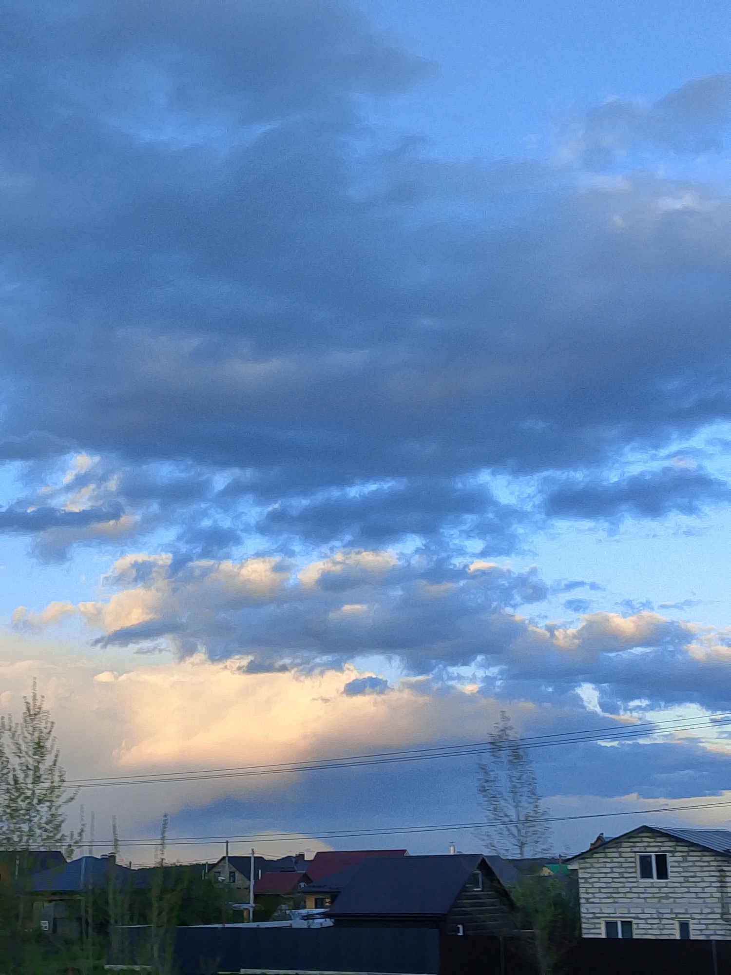 The image shows a cloudy sky above the rooftops of houses in Прикамский, Russia. The sky is dominated by dark blue and gray clouds, with patches of light blue visible. Some of the clouds near the horizon are illuminated with a warm, yellow light. Below the clouds, the rooftops of several houses are visible, varying in style and color, from dark roofs to lighter-colored walls. The image also contains silhouettes of trees and foliage along the bottom edge. Power lines run across the lower part of the sky in the middle ground.
