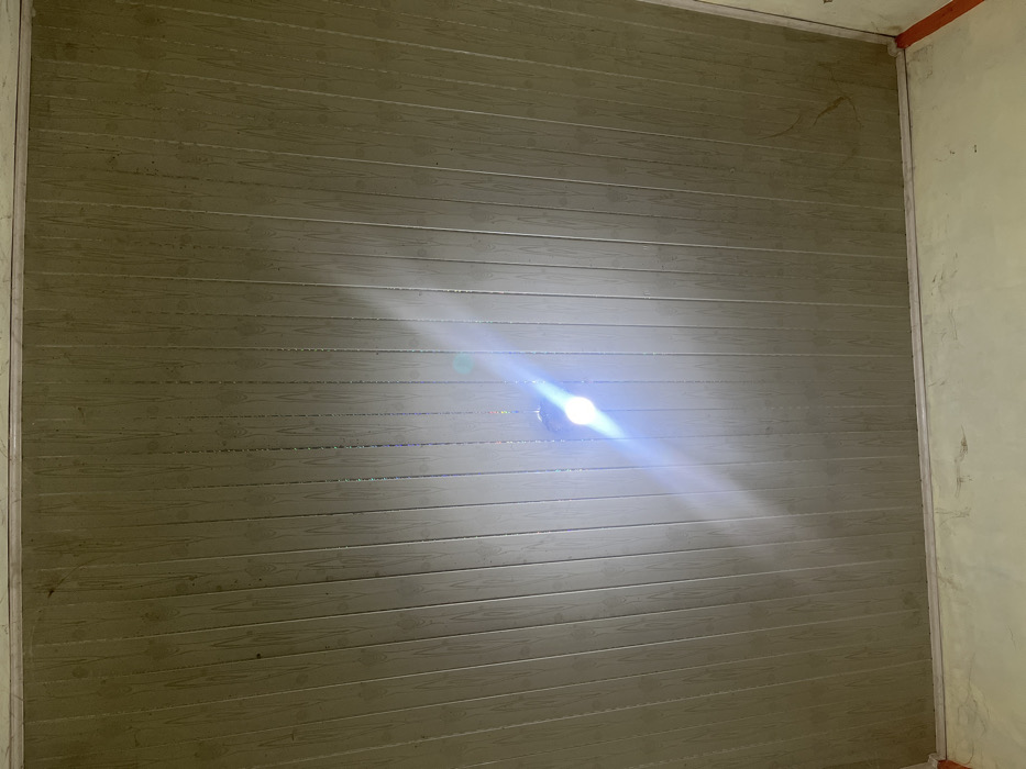 A textured wall with horizontal planks mimicking wood grain dominates the scene. A bright, diffused light source creates a strong glare near the center of the wall, obscuring the details in that area. The wall is framed by unfinished corners, suggesting ongoing construction or renovation. The adjacent walls appear unpainted or plastered, adding to the unfinished aesthetic.