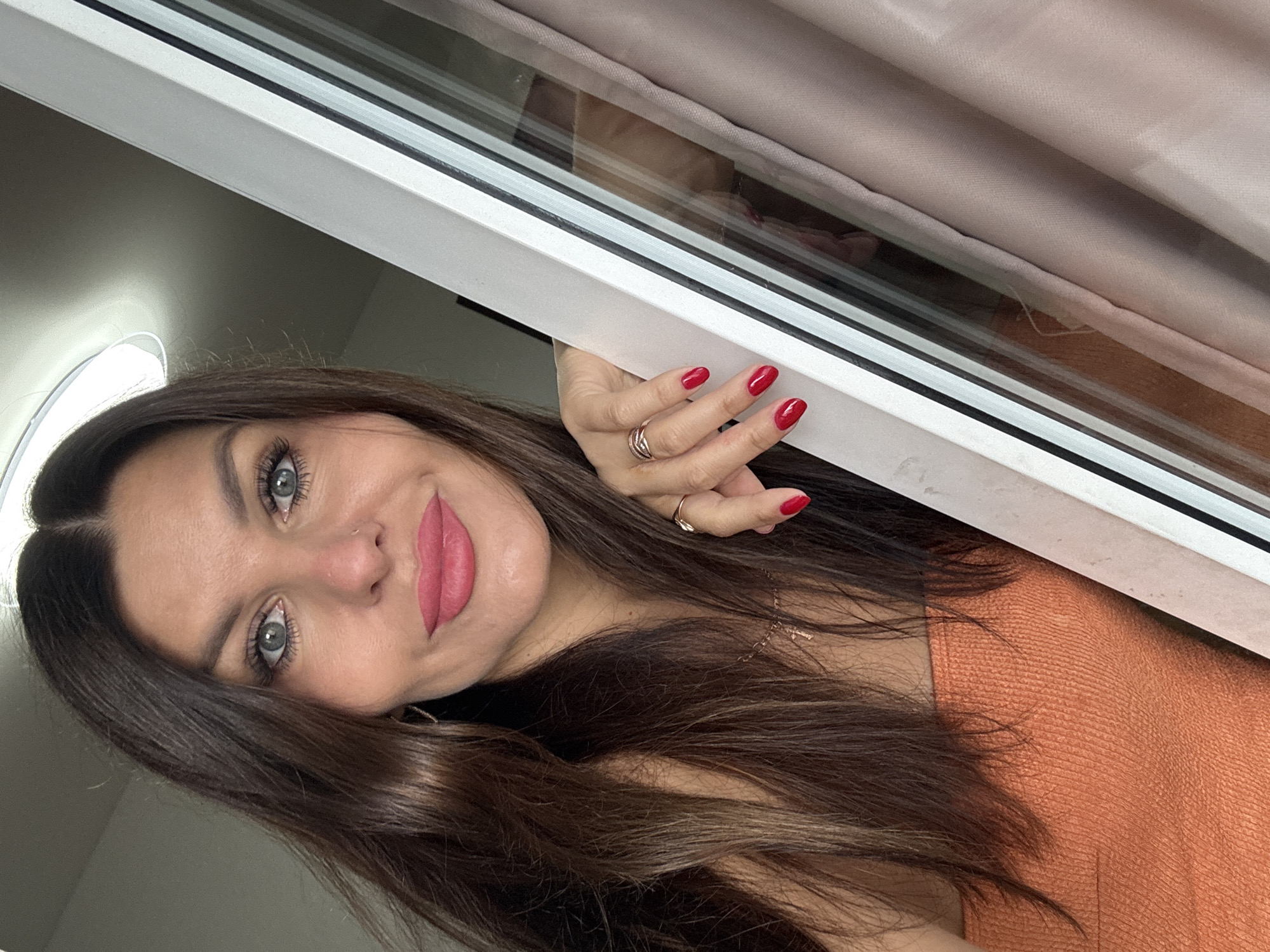 A woman with long, dark brown hair, blue-grey eyes, and bright pink lipstick leans towards the camera. She is wearing an orange strapless top and has red nail polish. Her right hand, adorned with rings, rests on a white frame. The background features a pale pink curtain and a window. This scene is not specifically located in Chaikovsky, Russia.