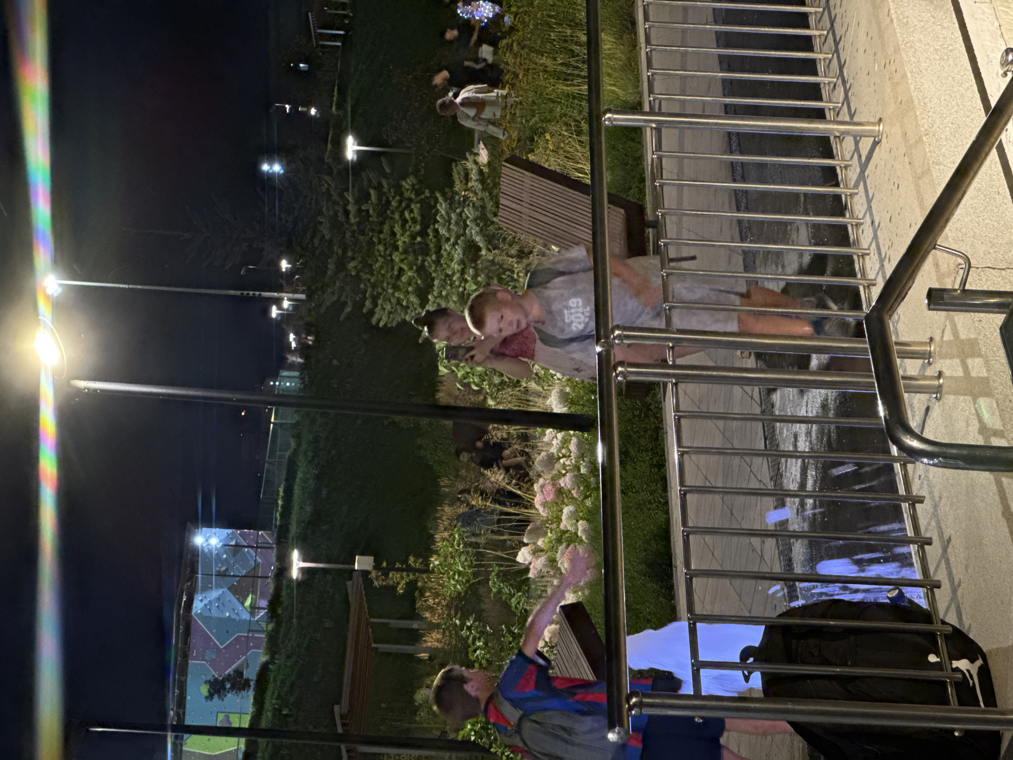 An outdoor scene at night, illuminated by streetlights and a brightly lit structure in the background, shows a group of people near a park-like area. In the foreground, a young boy wearing a blue and red striped shirt and a backpack is walking. To his right, another boy in a grey t-shirt with "2019" printed on it is also walking. Behind them, a woman is holding a phone and appears to be taking a photo, with other people visible in the mid-ground and background. A black backpack is placed on the ground near a metal railing. In the far background, a building with a colorful mural and climbing walls is visible, along with more trees and lights. The ground appears wet, suggesting recent rain or a water feature.