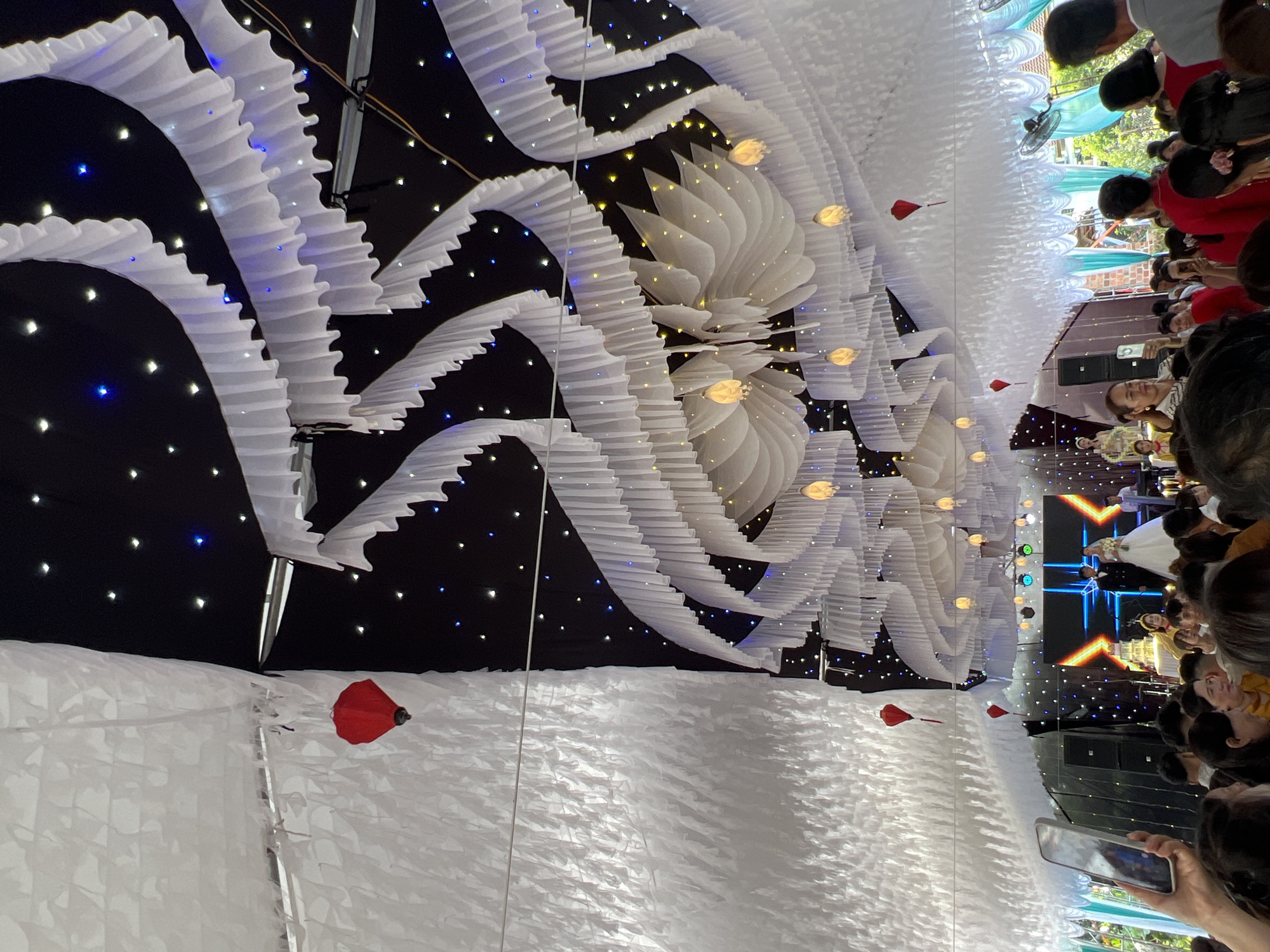 This image captures an indoor or covered event space, likely a festive celebration or reception. The ceiling is ornately decorated with flowing white fabric structures, resembling waves or stylized petals. These are interspersed with a black backdrop featuring numerous small twinkling lights in white, blue, and yellow, creating a 'starry night' ambiance. Several larger, warm-toned, lantern-like lights and small red heart-shaped or traditional lantern decorations are suspended from the ceiling.
In the foreground, a crowd of people, mostly viewed from behind, forms an audience, intently looking towards a stage. One person in the immediate foreground holds a smartphone, seemingly recording the event.
At the far end of the venue, a brightly lit stage is visible. A large LED screen on the stage displays abstract blue and orange geometric patterns. On the stage, a man in a suit and a woman in a white gown, possibly a wedding dress, are present, alongside another individual in a yellow traditional outfit. Speakers and stage lighting are also visible.
The scene suggests an evening event, possibly a wedding reception, given the attire of individuals on stage and the celebratory decorations. The event is taking place in Bình An, Vietnam. No discernible text is visible.