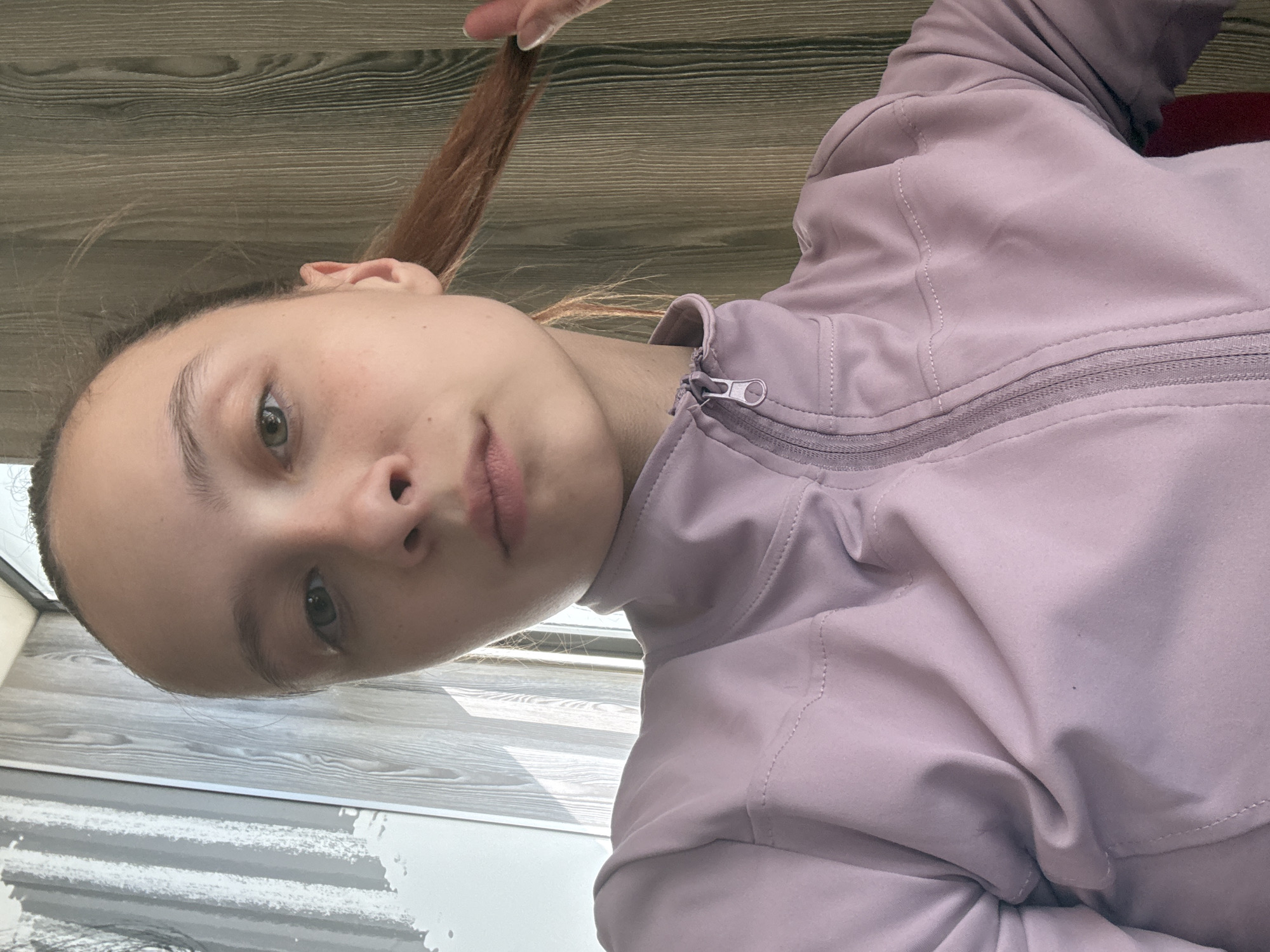 A close-up, angled shot of a person with fair skin, light brown hair held up by a hand, and green eyes, wearing a light purple zip-up jacket. The background consists of a wooden surface and a section of tiled flooring with a gray and white pattern. The person appears to be indoors, with the lighting soft and diffused. There is no information available about the city of Perm, Russia, in the image.