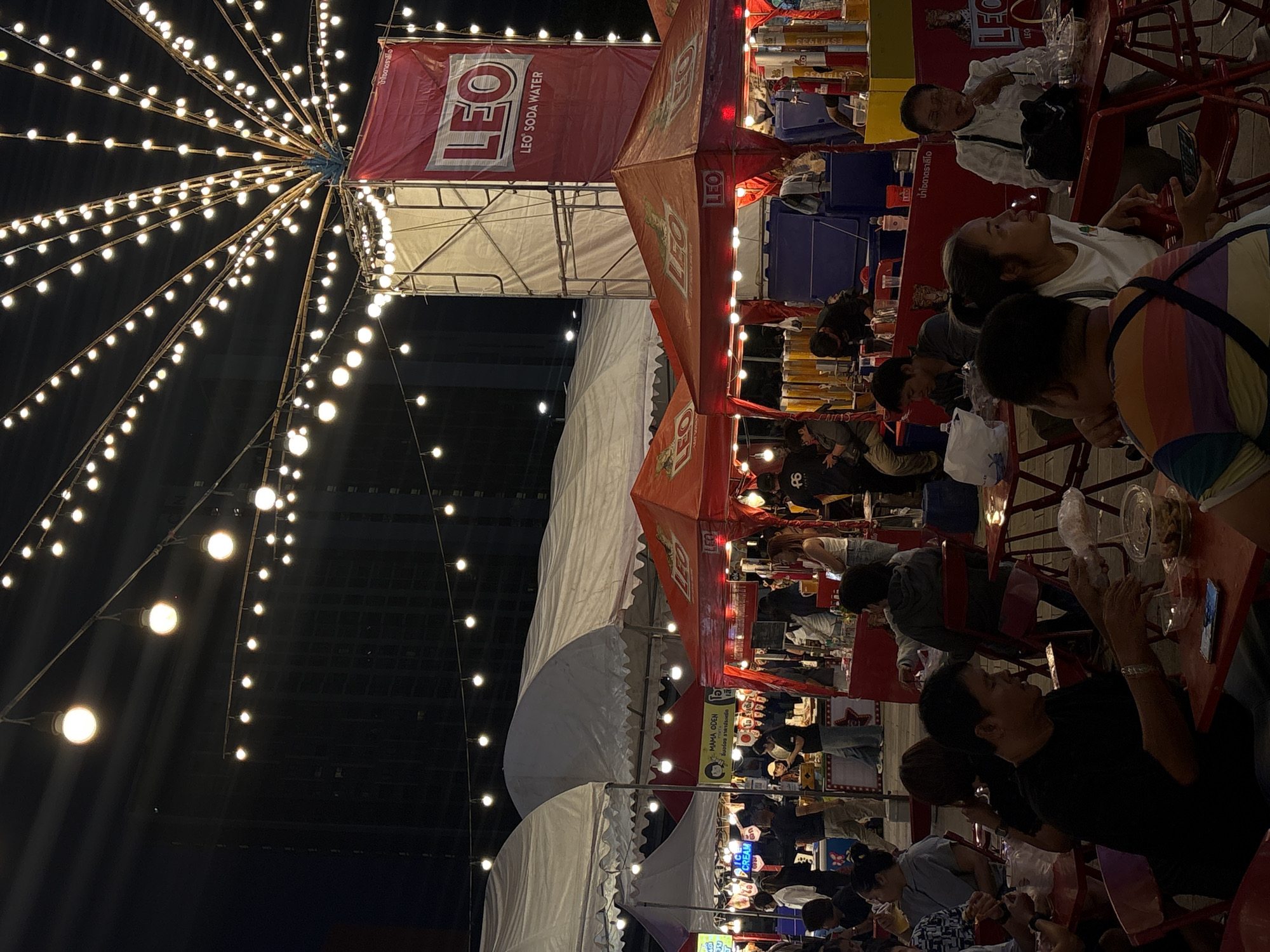 An outdoor night market in Chiang Mai, Thailand, is depicted. The scene is illuminated by numerous strings of bright, exposed light bulbs that hang from a central structure and extend outwards like a fan. In the background, a tall, dark building is visible, its windows reflecting the light. Several market stalls with red and white awnings are set up. One prominent stall features a large red banner with the "LEO" logo and the text "LEO SODA WATER." Another stall has a sign that says "MAMA ODEN" and "ICE CREAM." Numerous people are present, some standing and others seated at red tables, eating and socializing. The foreground shows several individuals seated at tables, with food and drinks laid out before them. The atmosphere is lively and suggests a typical evening at a bustling night market.