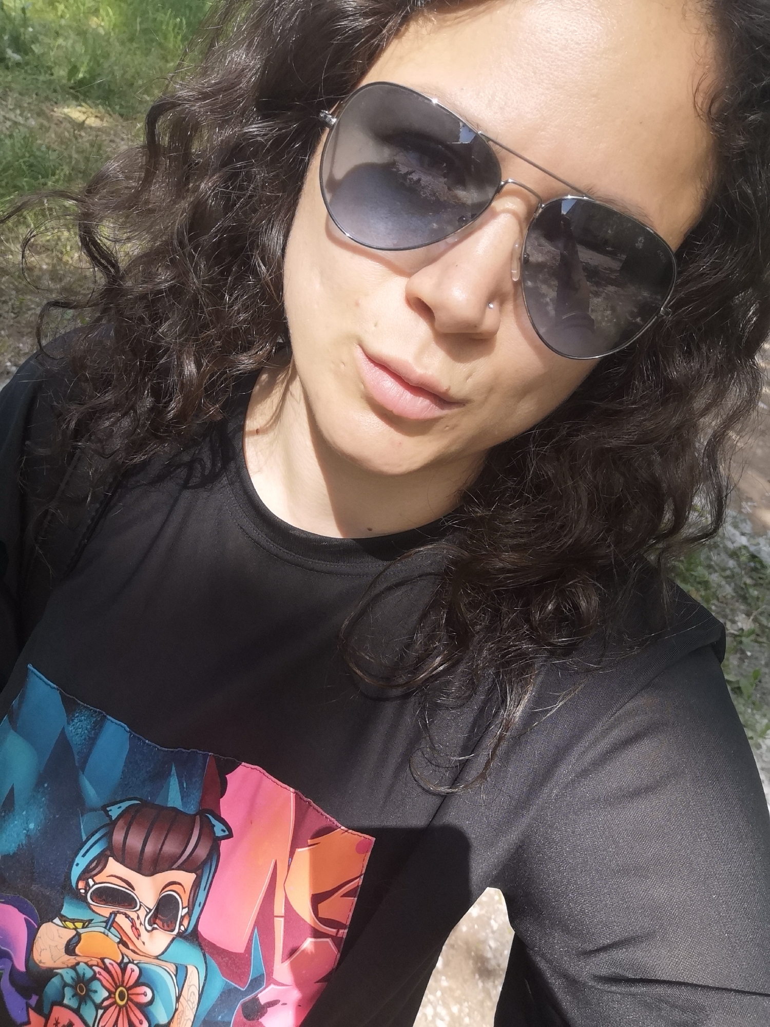 A woman is taking a selfie outdoors. She has dark, curly hair, and is wearing aviator sunglasses, a black t-shirt with a colorful graphic print, and a nose ring. The background appears to be a grassy area with some dirt and sunlight dappling the scene. The specific location in Chaikovsky, Russia, cannot be determined from the image.