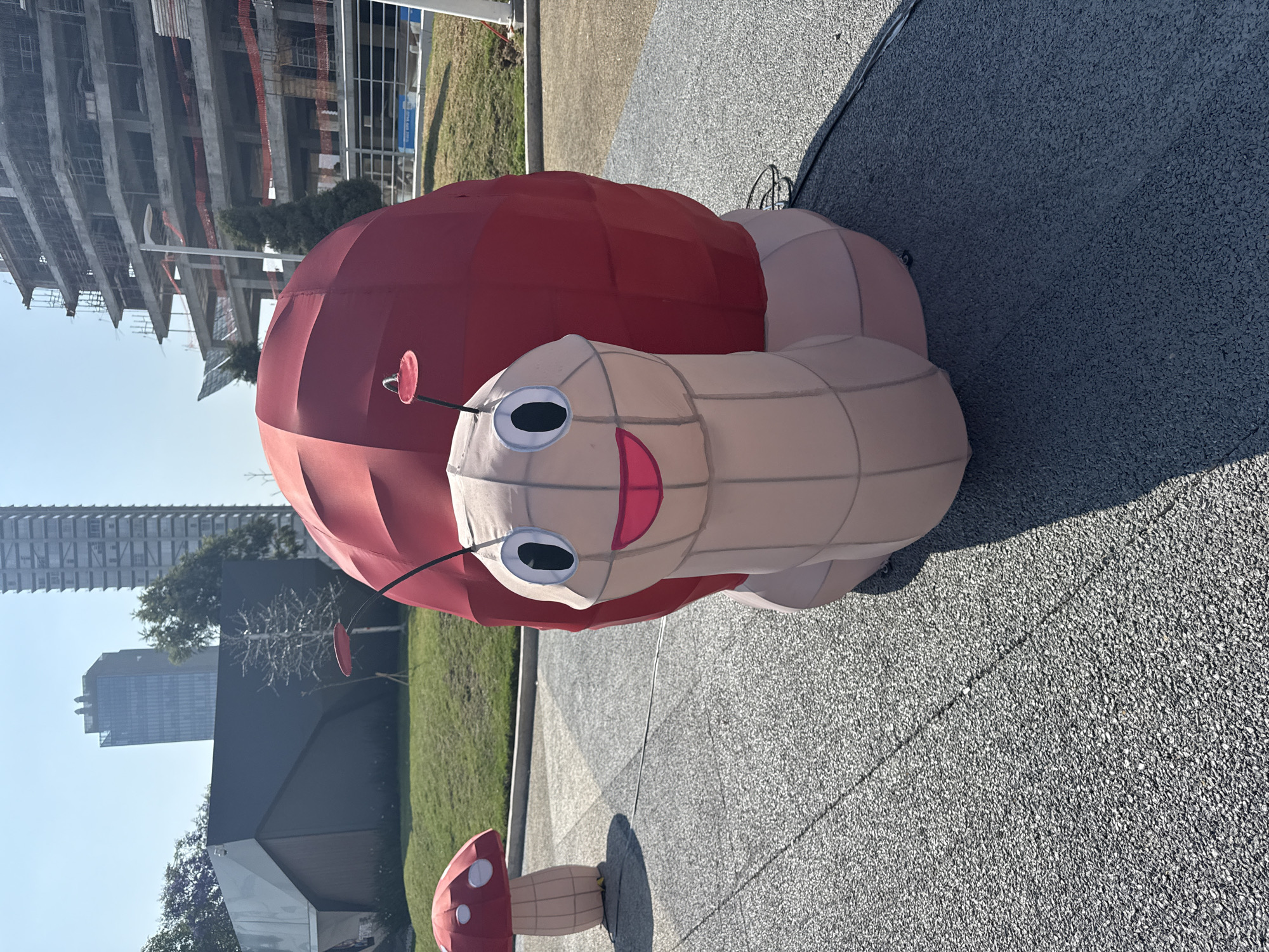 A large, red and pink snail sculpture with cartoonish eyes and a smiling mouth is displayed on a gray stone surface. A smaller mushroom sculpture is also visible nearby. Tall buildings and construction are visible in the background. The photograph appears to be taken outdoors in Puebla, Mexico.