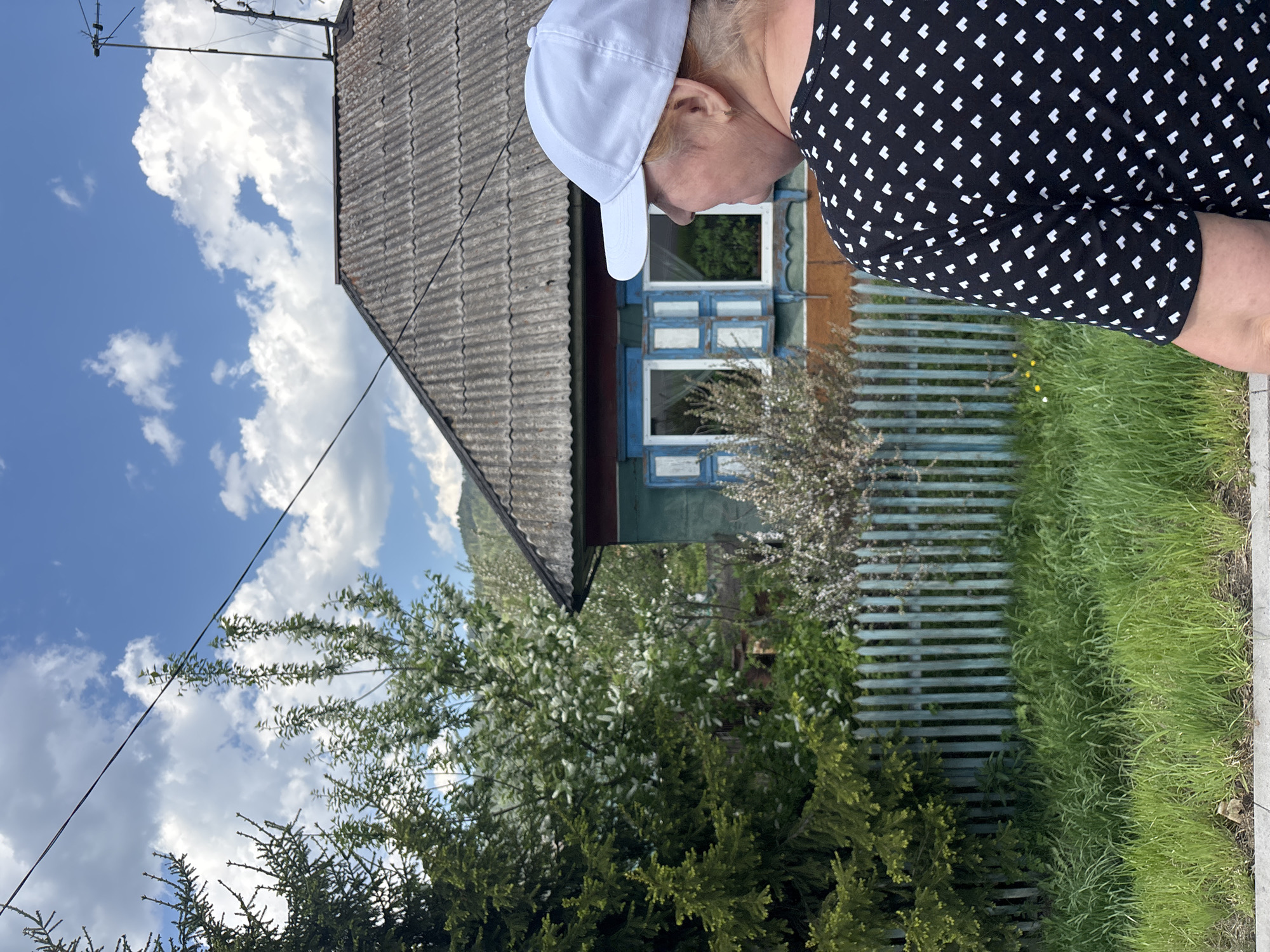 A medium shot captures a woman in a black and white patterned dress and white baseball cap standing next to a small, one-story house. The house has a gray, corrugated roof and blue window frames. A wooden fence in a matching blue color runs along the side of the house, bordered by grass. Lush green bushes and trees fill the foreground, with a sky full of white clouds visible behind the house. This location is Овсянка, Russia.