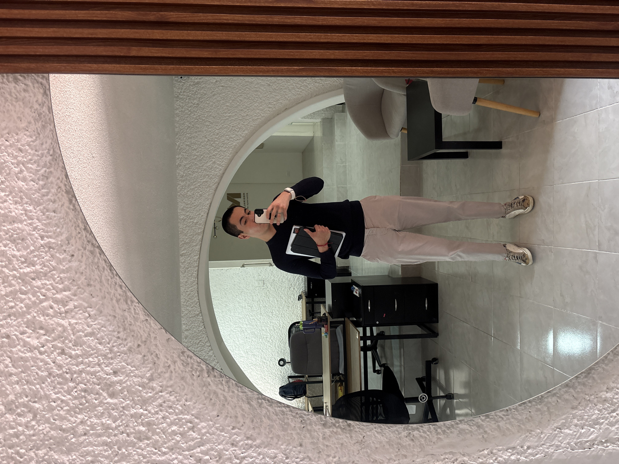 A medium shot captures the reflection in a round mirror with a textured, off-white frame. The mirror also has a decorative wooden top. A man is reflected, holding a smartphone and a tablet. He is wearing a black long-sleeved shirt and light-colored pants with white sneakers.
The background, visible through the mirror, is an office space with tiled floors. Office furniture, including a desk, black cabinets, and chairs, is visible. In the distance, a waiting area with gray chairs and a black table can be seen.
The office is located in Puebla, Mexico.