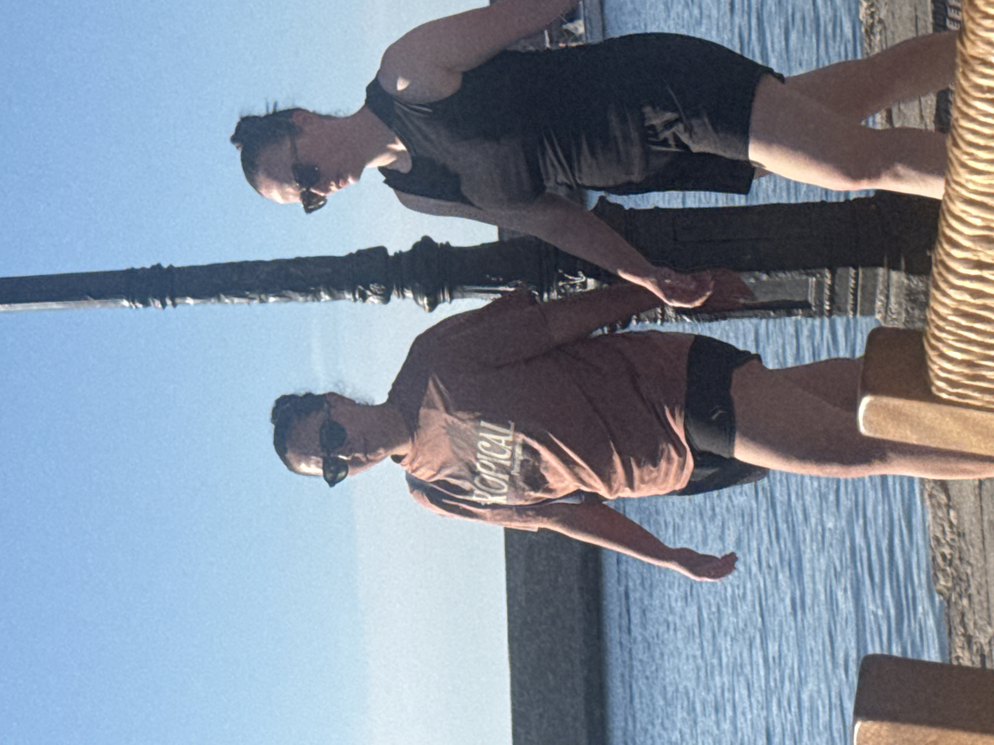 Two women walk hand-in-hand along a waterfront promenade in Chania, Greece. The woman on the left wears a coral-colored t-shirt with the word "TROPICAL" printed on it in white text, and black shorts. She has sunglasses on and her hair is pulled up in a bun. The woman on the right wears a black sleeveless top and black shorts. She also has sunglasses on and her hair is in a bun. They are walking past a black lamppost. The sea is to their left, with a stone wall and buildings in the background. The sky is a clear blue. In the foreground, there are parts of what appear to be wicker chairs.